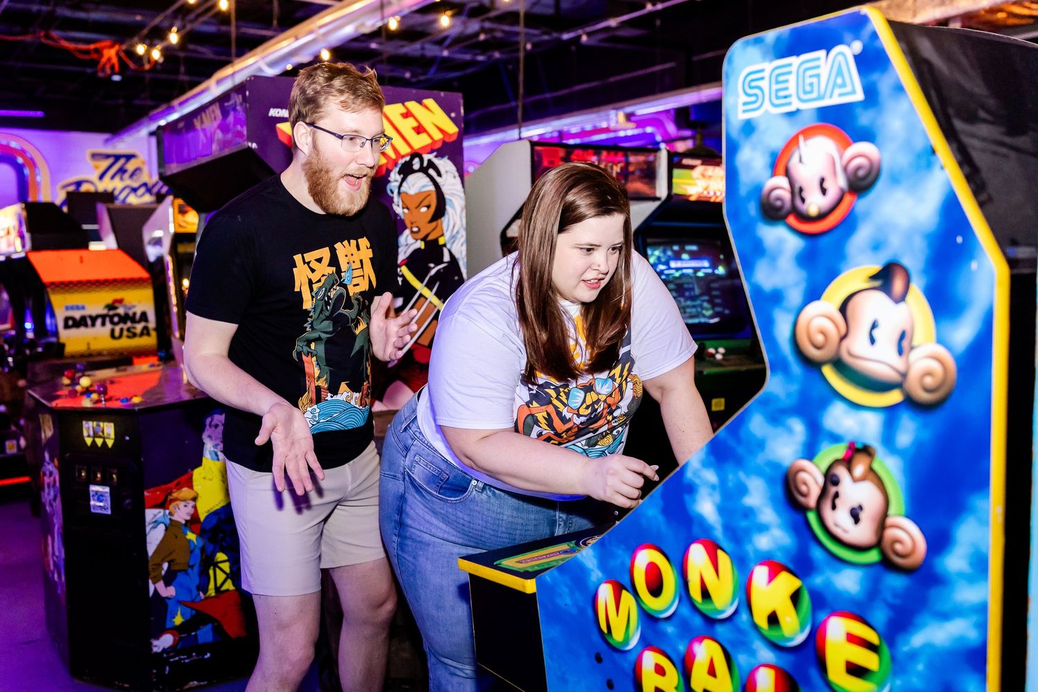 Two people playing arcade games at a neon-lit gaming venue near a Monkey Ball arcade cabinet.