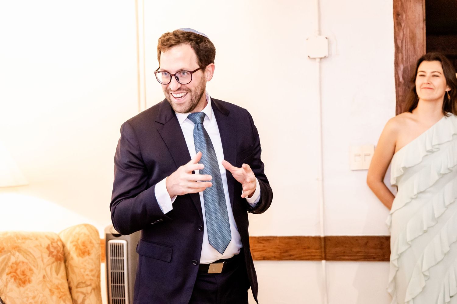 A person in formal attire gestures while speaking at an indoor wedding event.