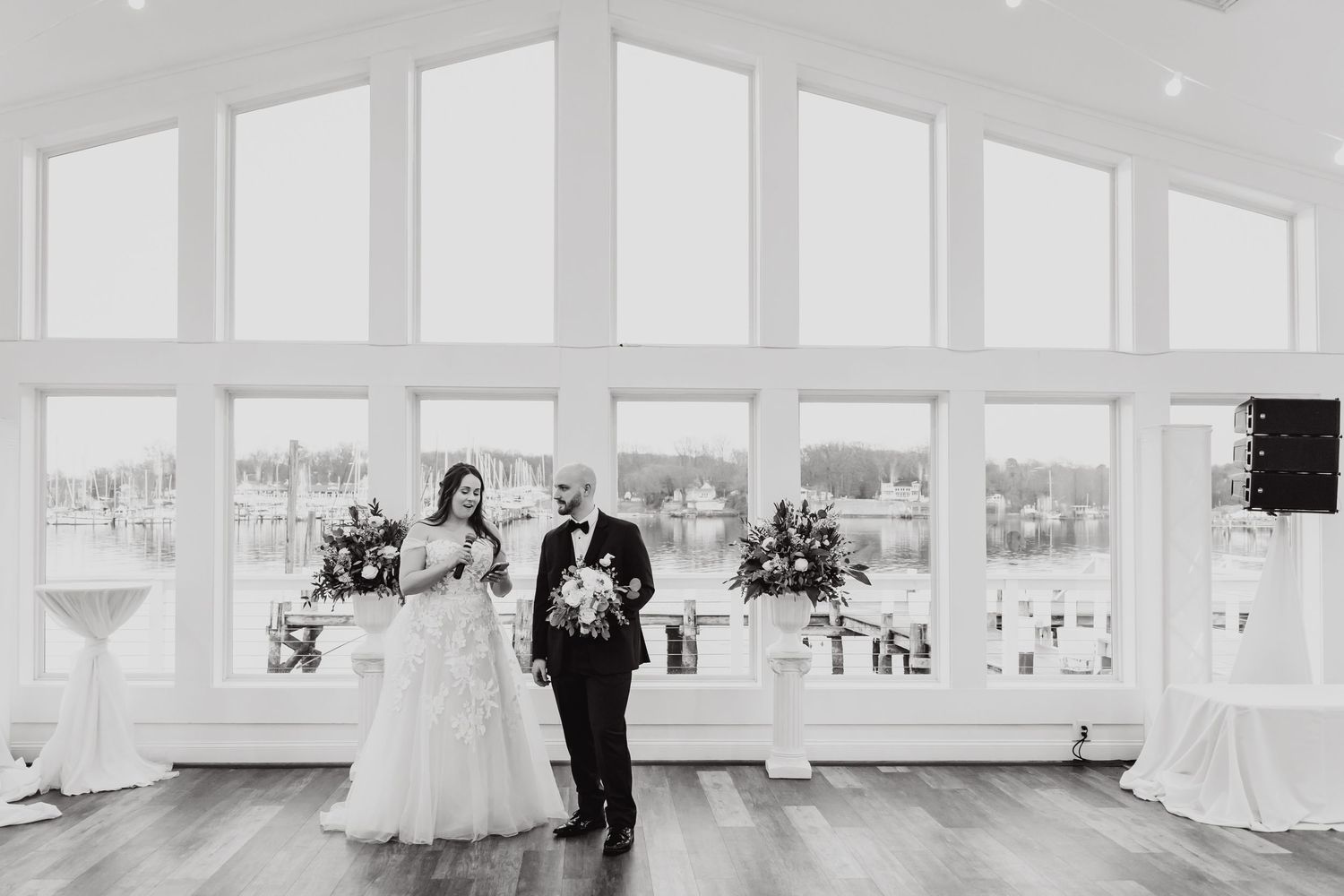 A black and white photograph of a wedding couple standing in an elegant room with large windows.