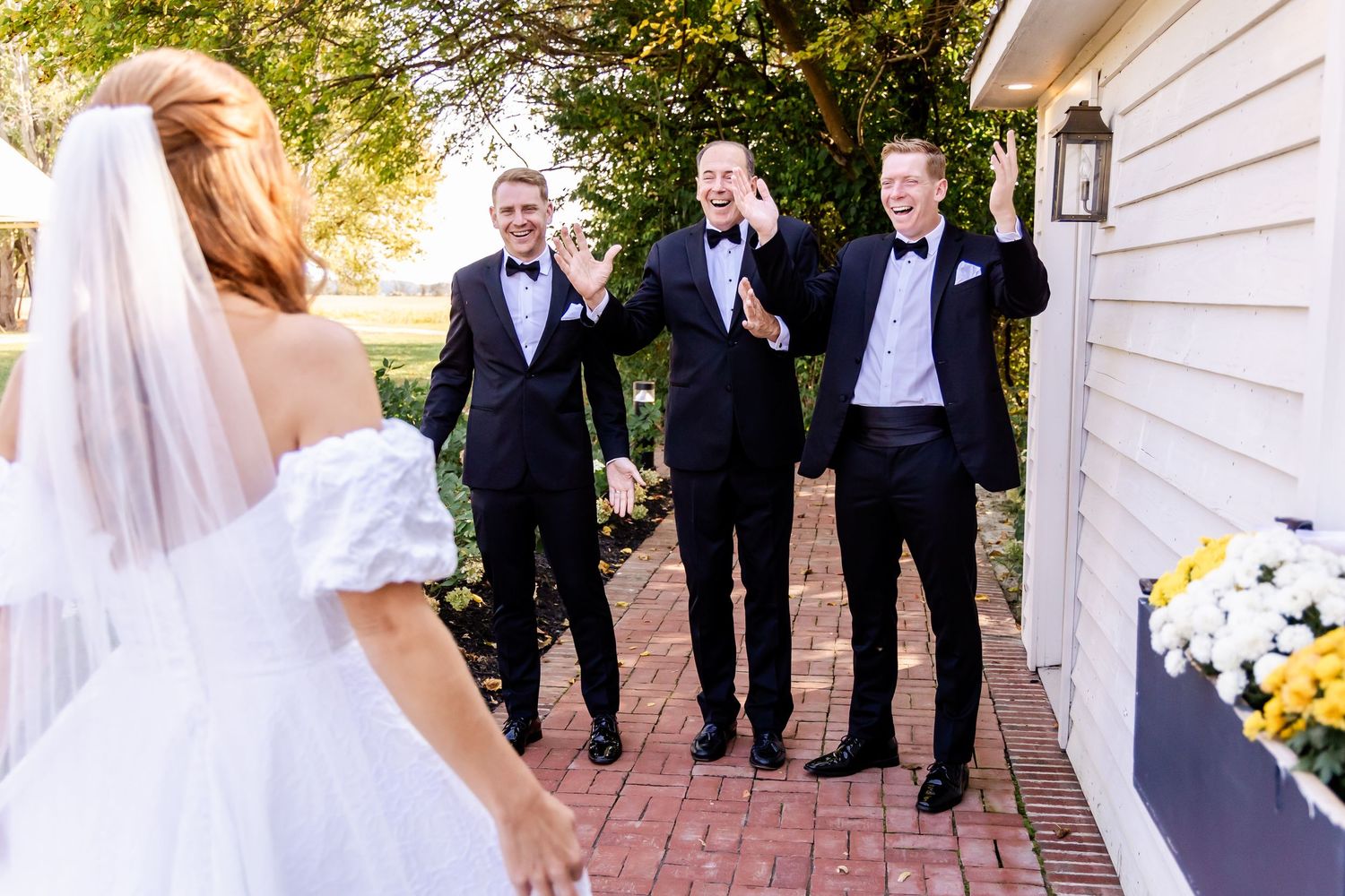 Groomsmen in black tuxedos react with excitement while waiting on brick path by white building.