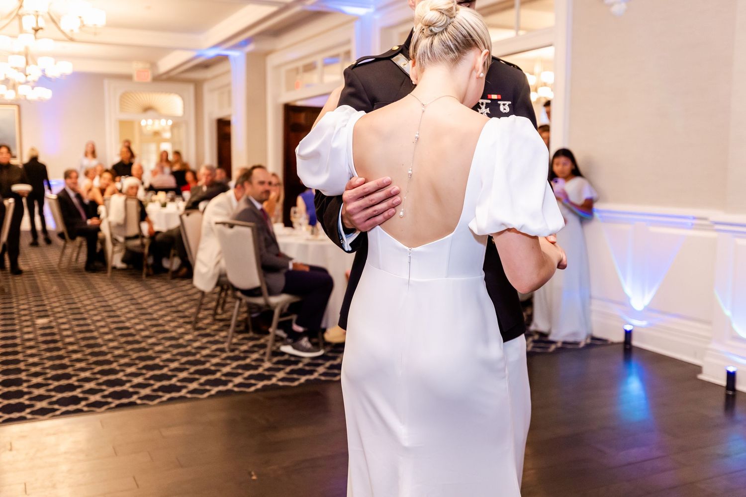 A wedding reception features couples dancing on a checkerboard floor beneath elegant chandeliers in a white ballroom.