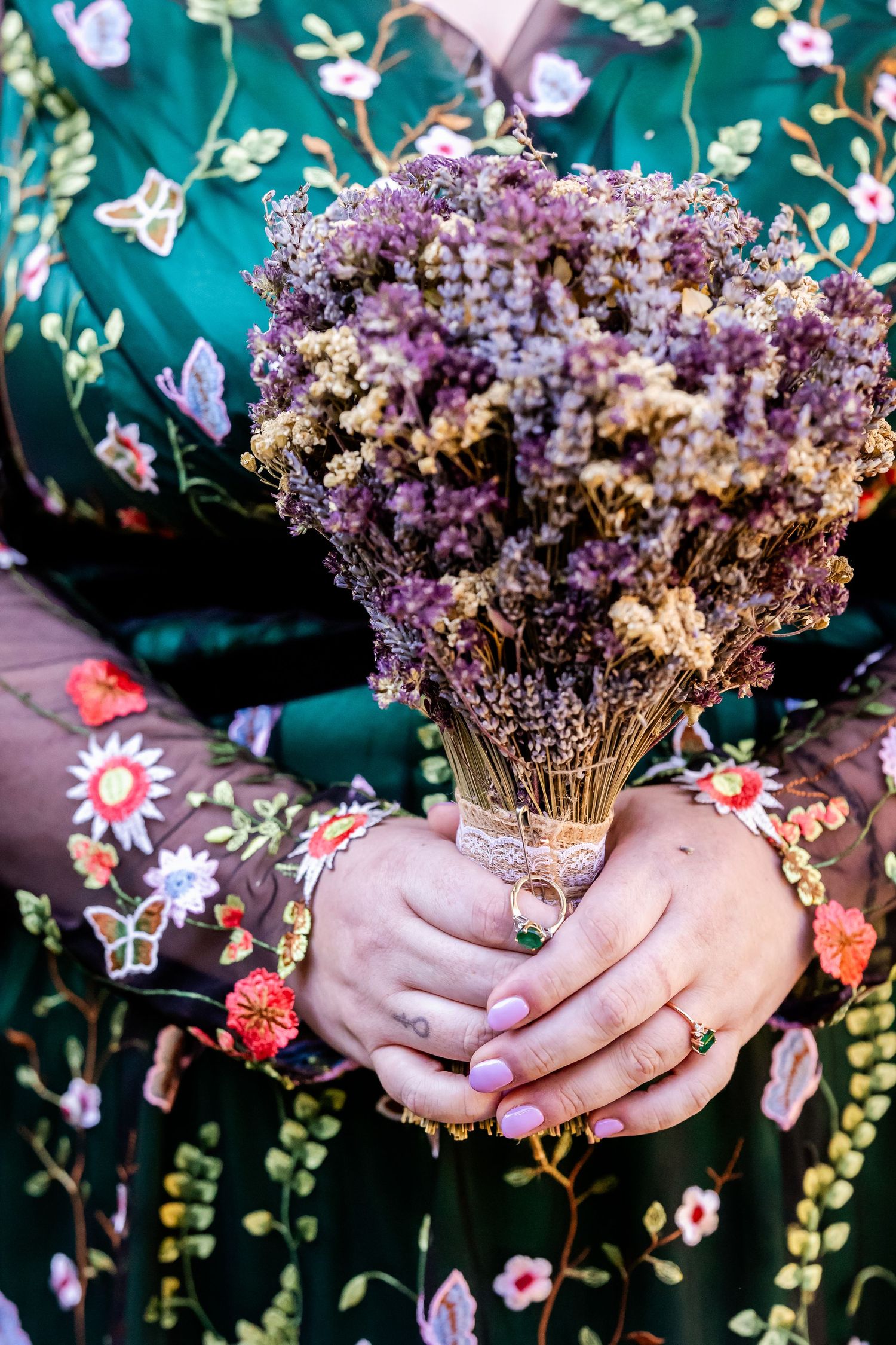 Hands holding a dried purple floral bouquet against a green floral print dress with delicate rings on fingers.