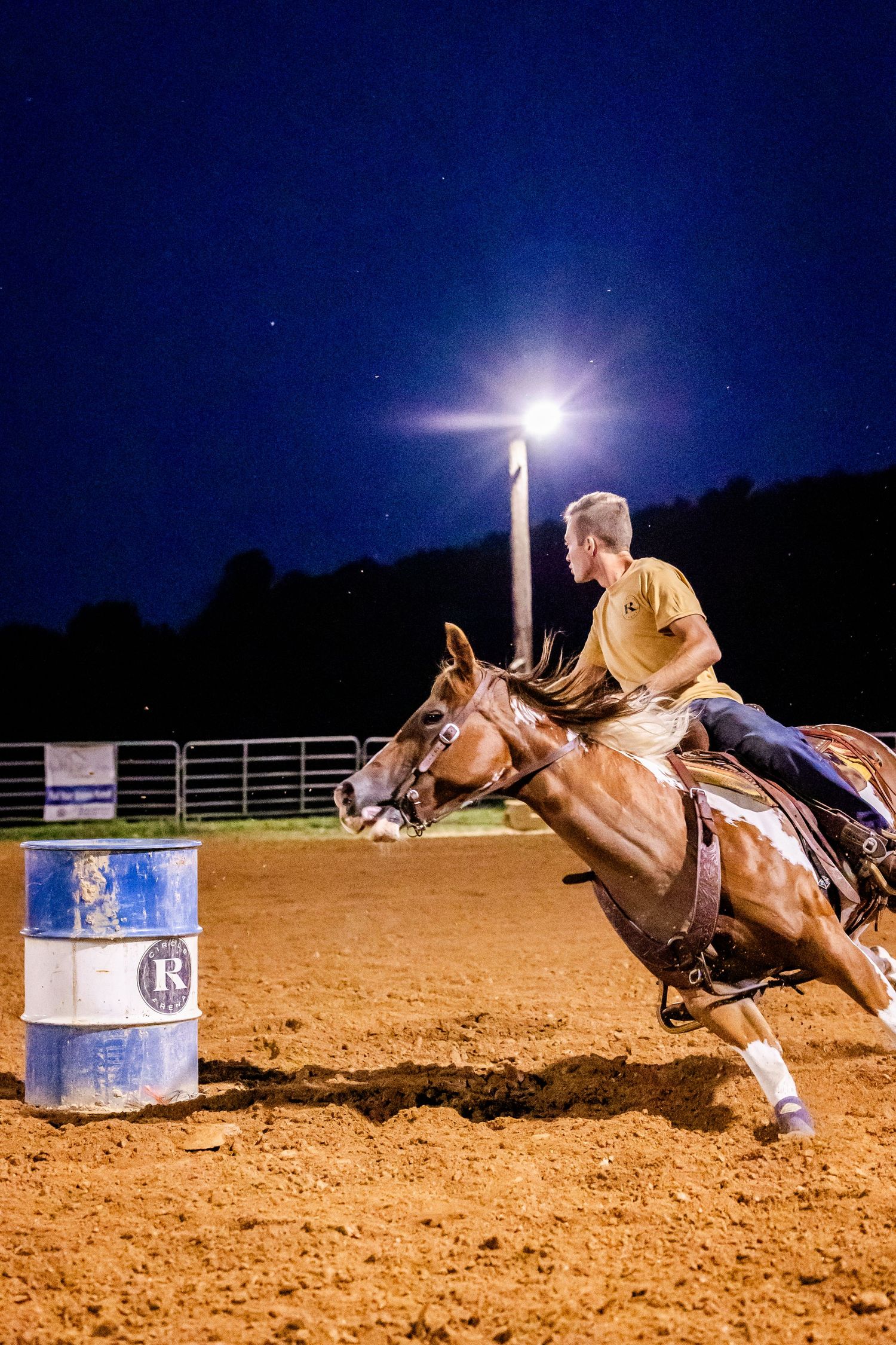 Barrel racing sequence at night showing horse and rider making a tight turn around blue metal barrel in sandy arena.