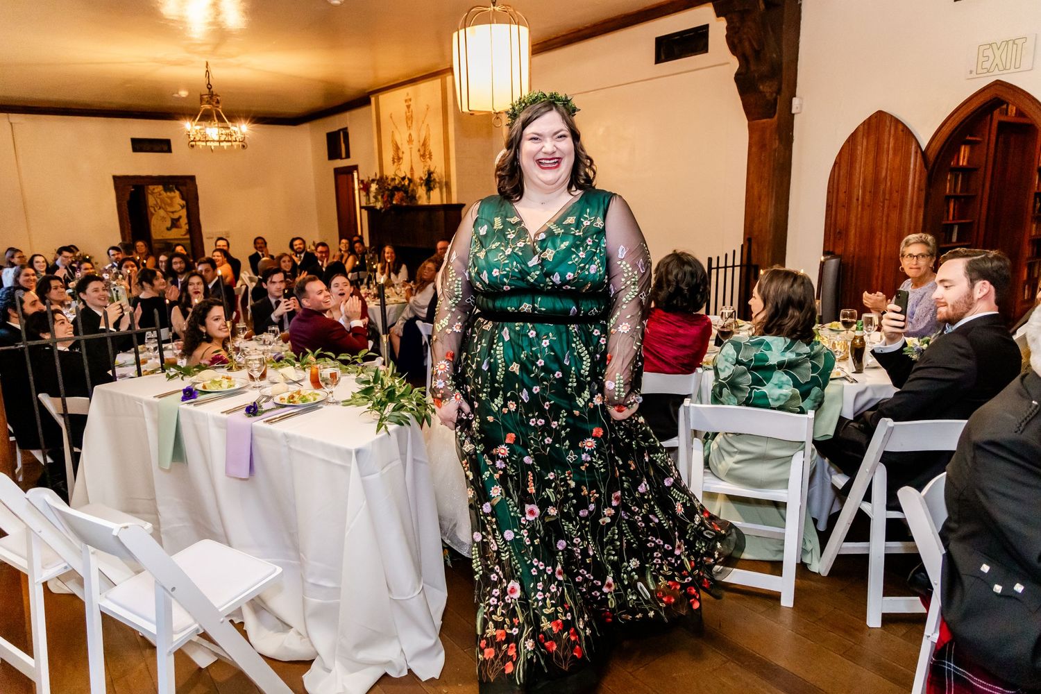 Woman in a green floral dress speaks at an elegant wedding reception with chandelier lighting.