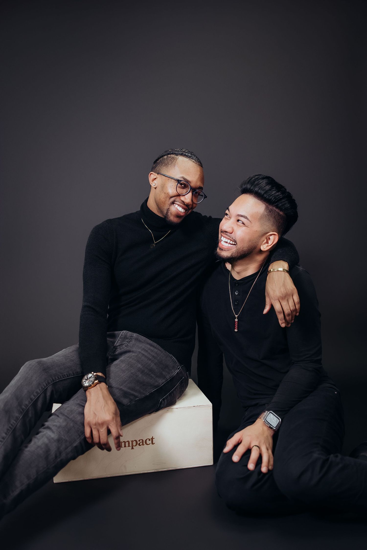 Two people in black clothing sharing candid moments on white bench against dark backdrop.