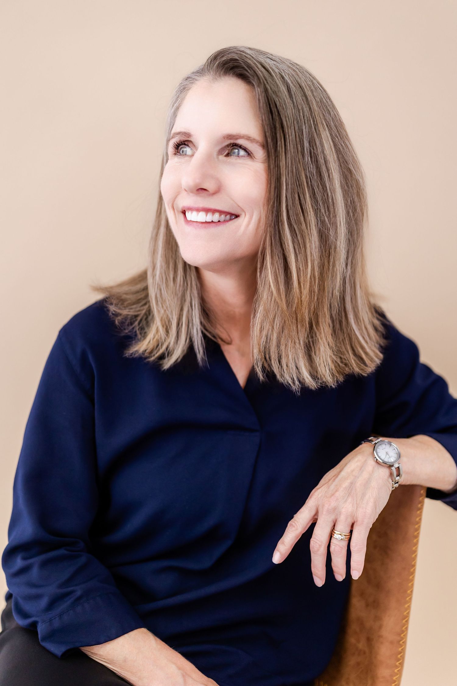 Smiling middle-aged woman in navy blouse sitting in chair, looking up, wearing a watch, against beige background.