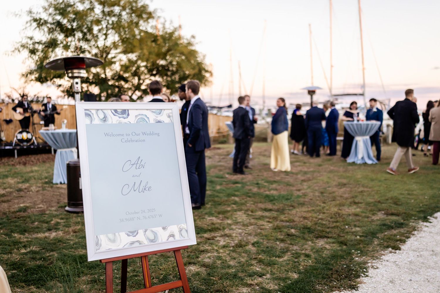 Elegant outdoor wedding welcome sign on easel with sailboats and guests mingling at waterfront venue at sunset.