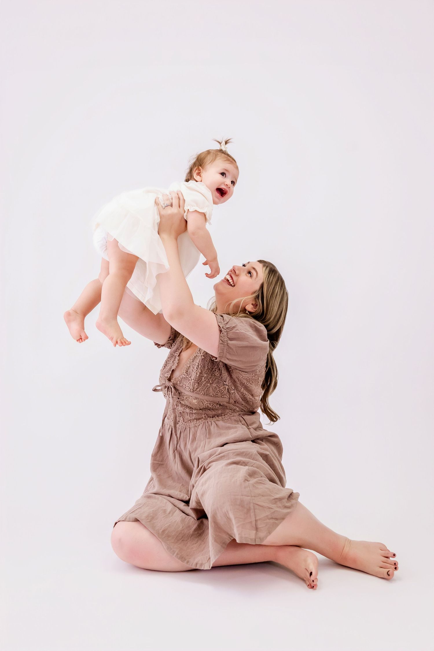 A parent in a beige dress plays lovingly with a baby in white against a light studio background.