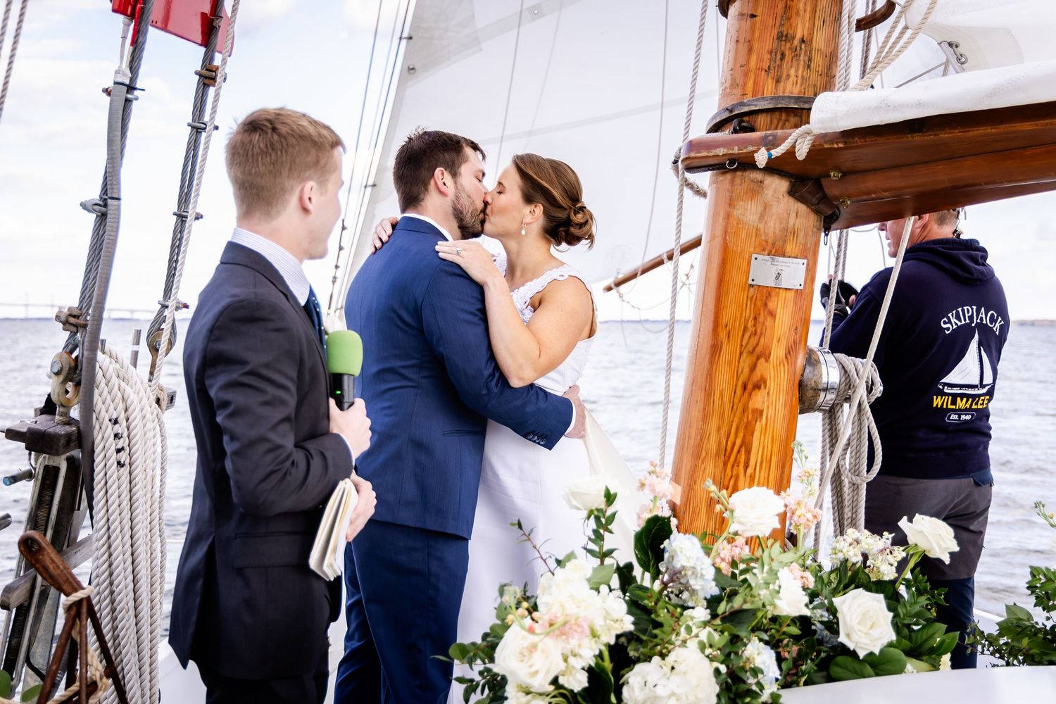 Bride and groom share a kiss on a sailboat deck during their wedding ceremony, surrounded by white floral arrangements.