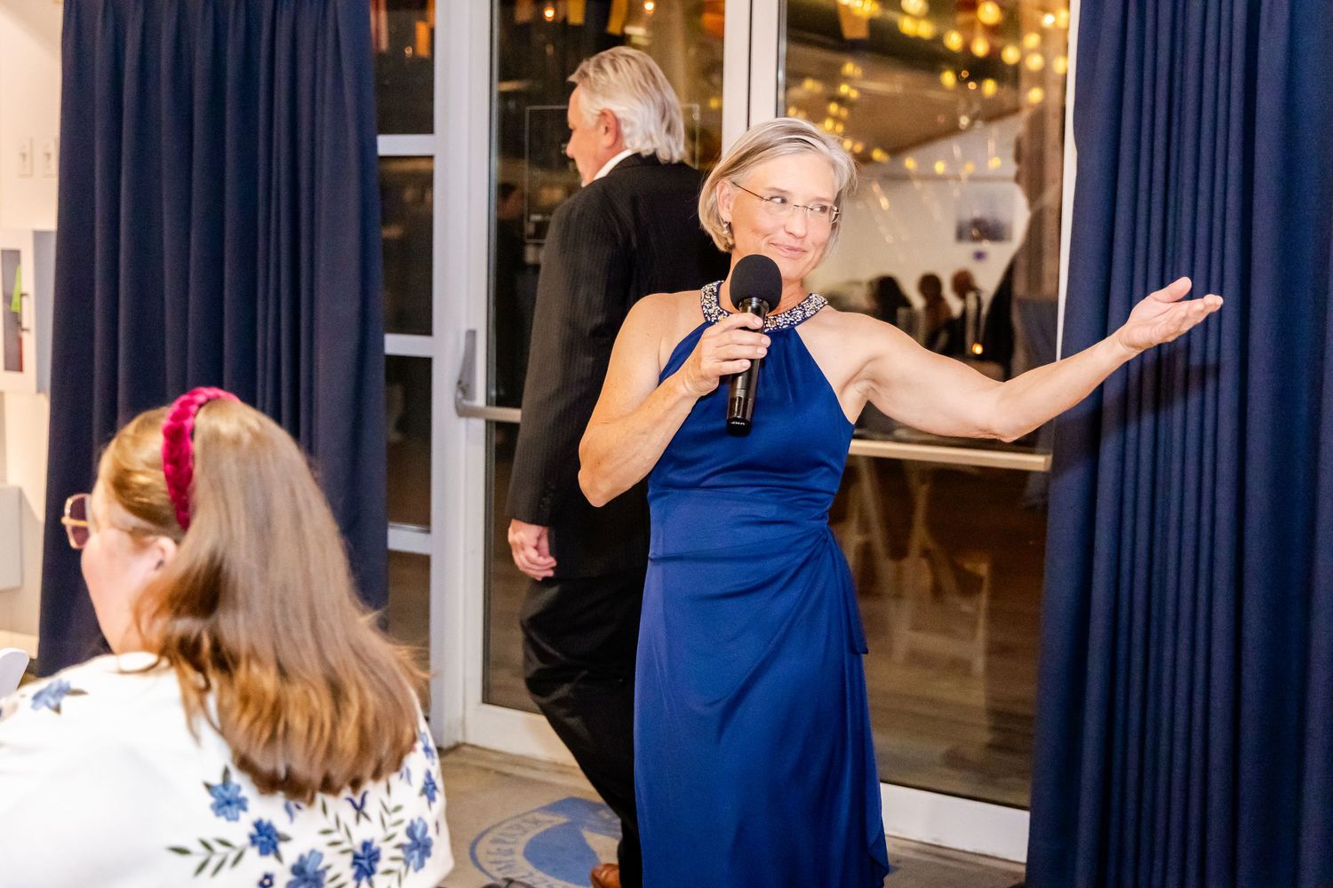 Woman in blue gown speaks into microphone at elegant evening event with guests seated nearby.