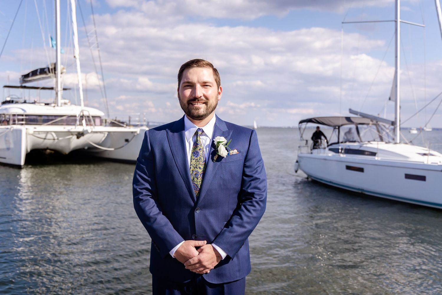 Groom in blue suit with floral tie poses at marina with sailboats on calm water under cloudy sky.