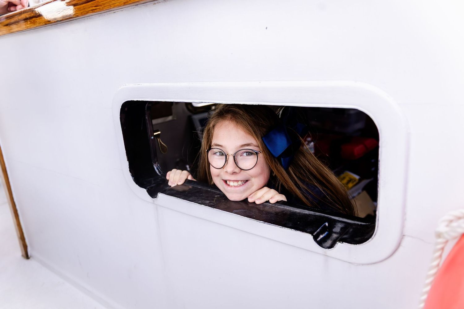 Young girl with glasses smiling through a small rectangular porthole window on a white boat.