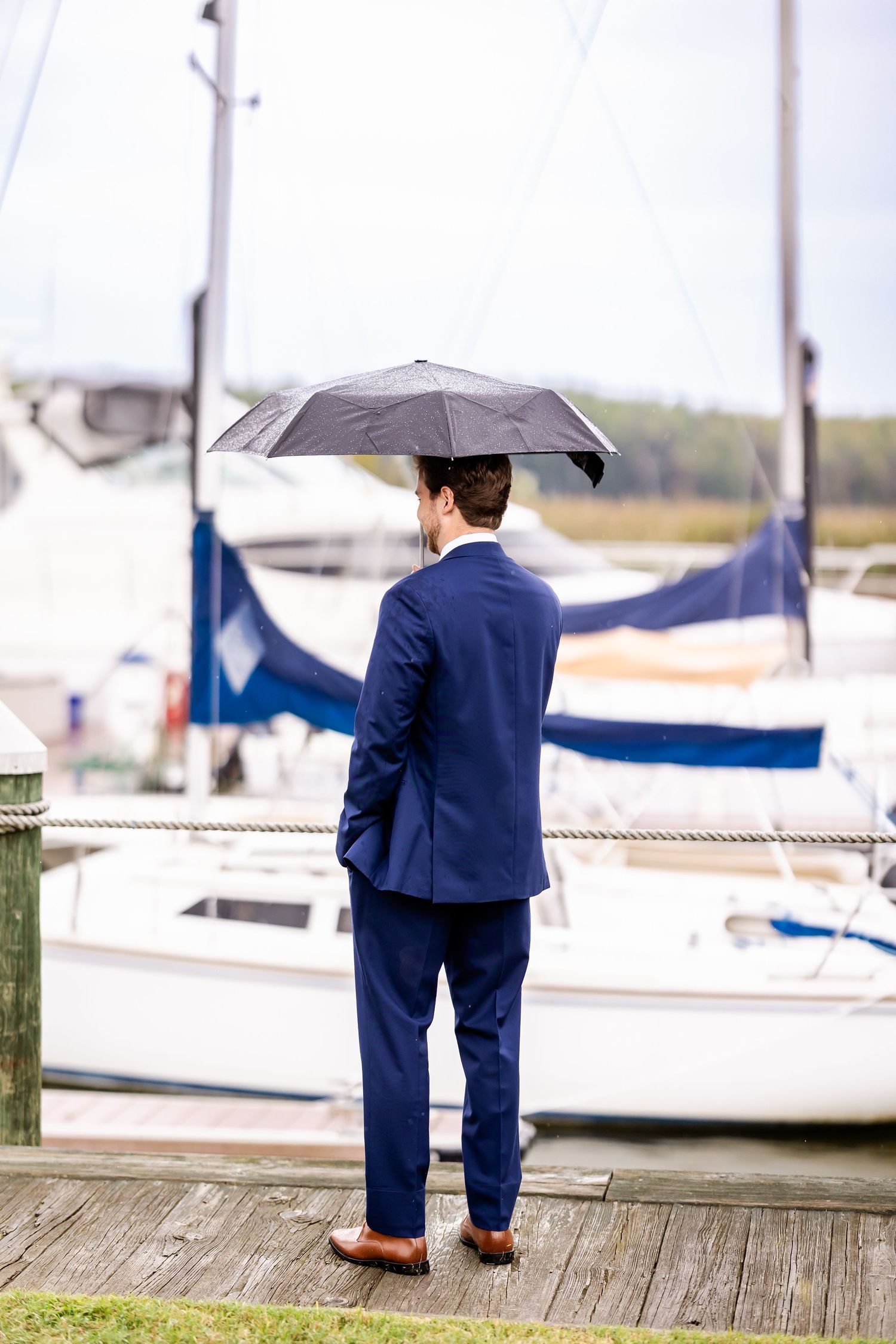 A person in a navy suit stands on a wooden dock overlooking boats and water on an overcast day.