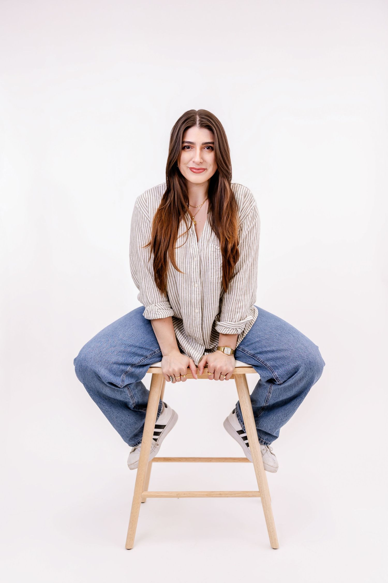 Young woman in striped shirt and jeans sitting on wooden stool against white background.