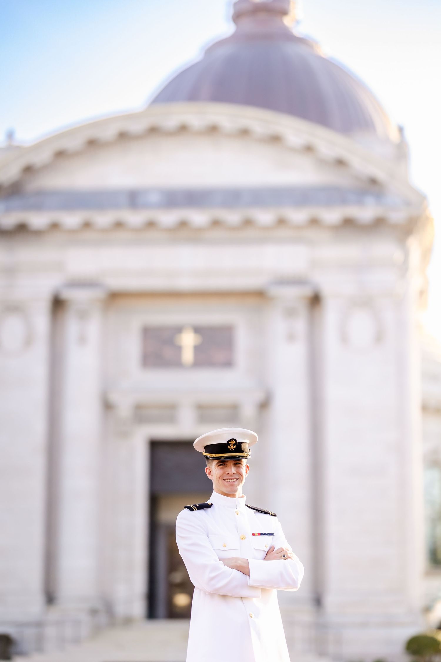 Naval officer in white dress uniform stands with arms crossed before a grand domed chapel building.