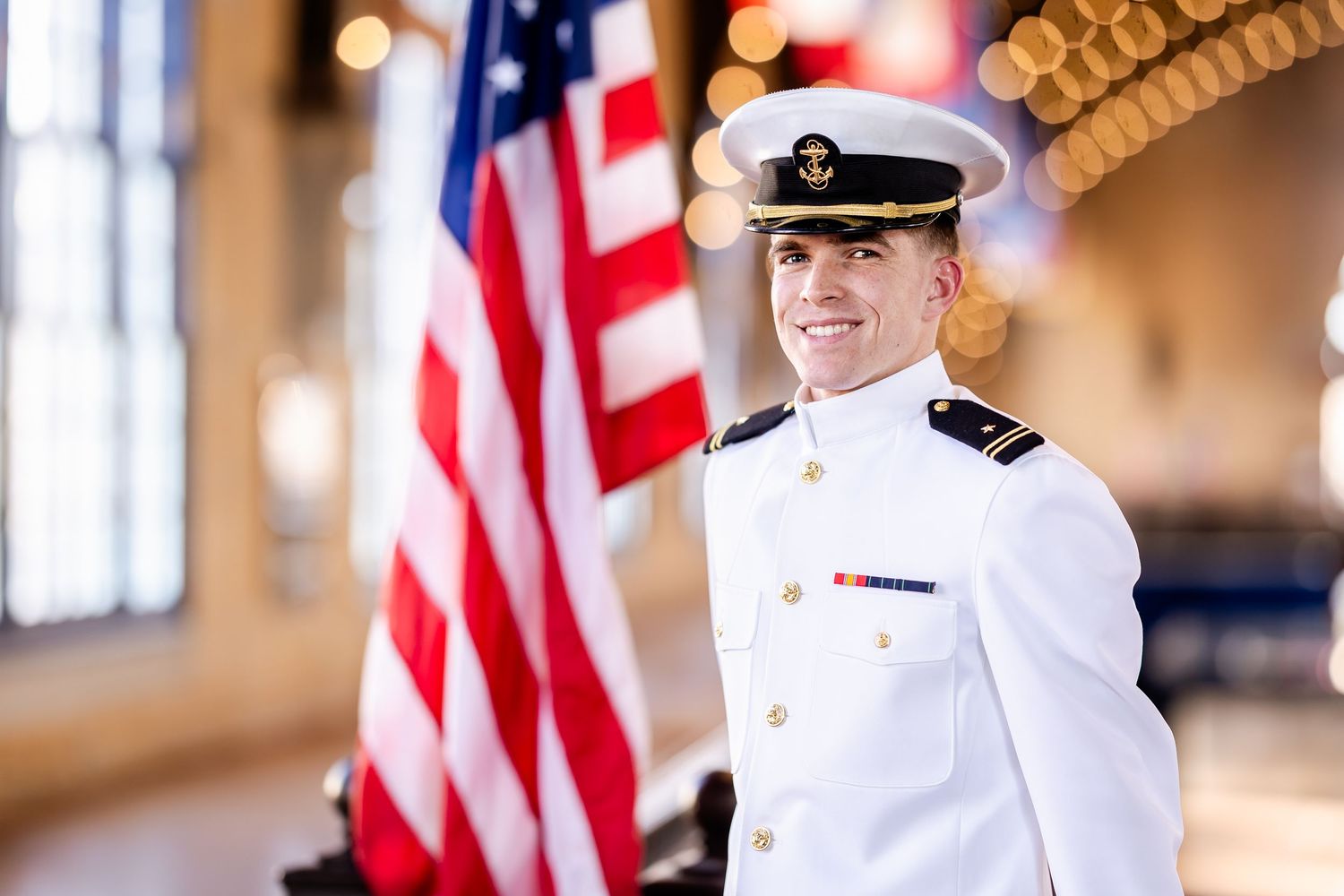 Naval officer in white dress uniform smiling proudly before an American flag in a formal indoor setting.