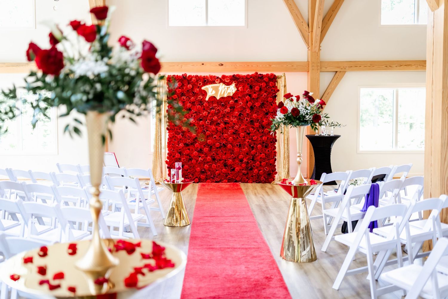 Elegant wedding venue with red rose wall, gold vases, and white chairs lining red carpet aisle.