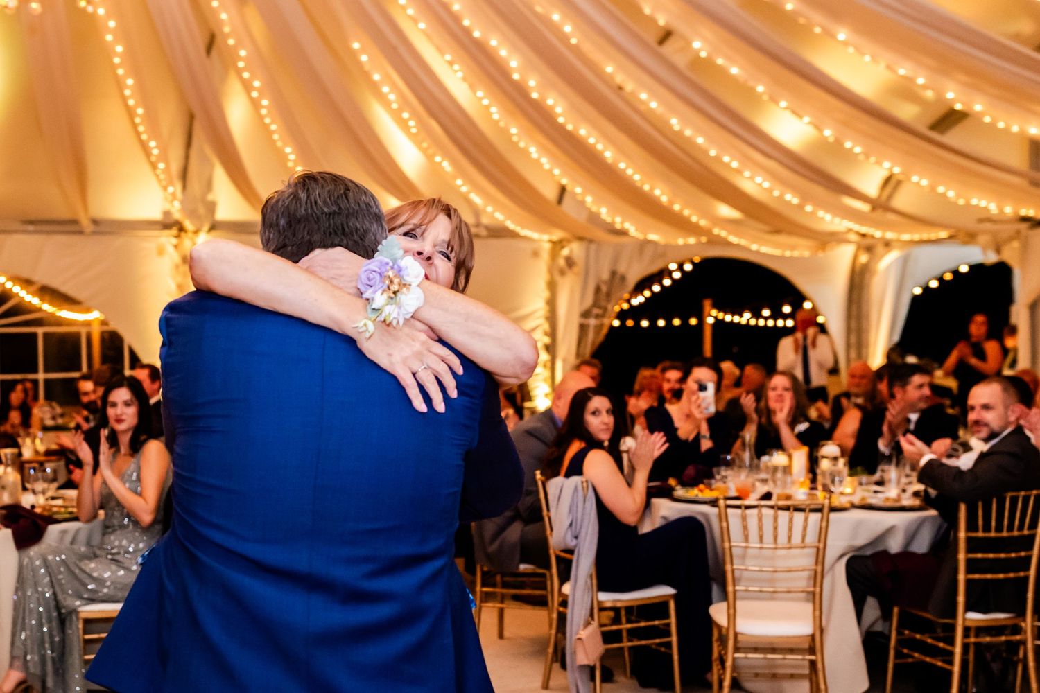 Couple shares an emotional dance under a beautifully lit tent ceiling with guests watching at their reception.