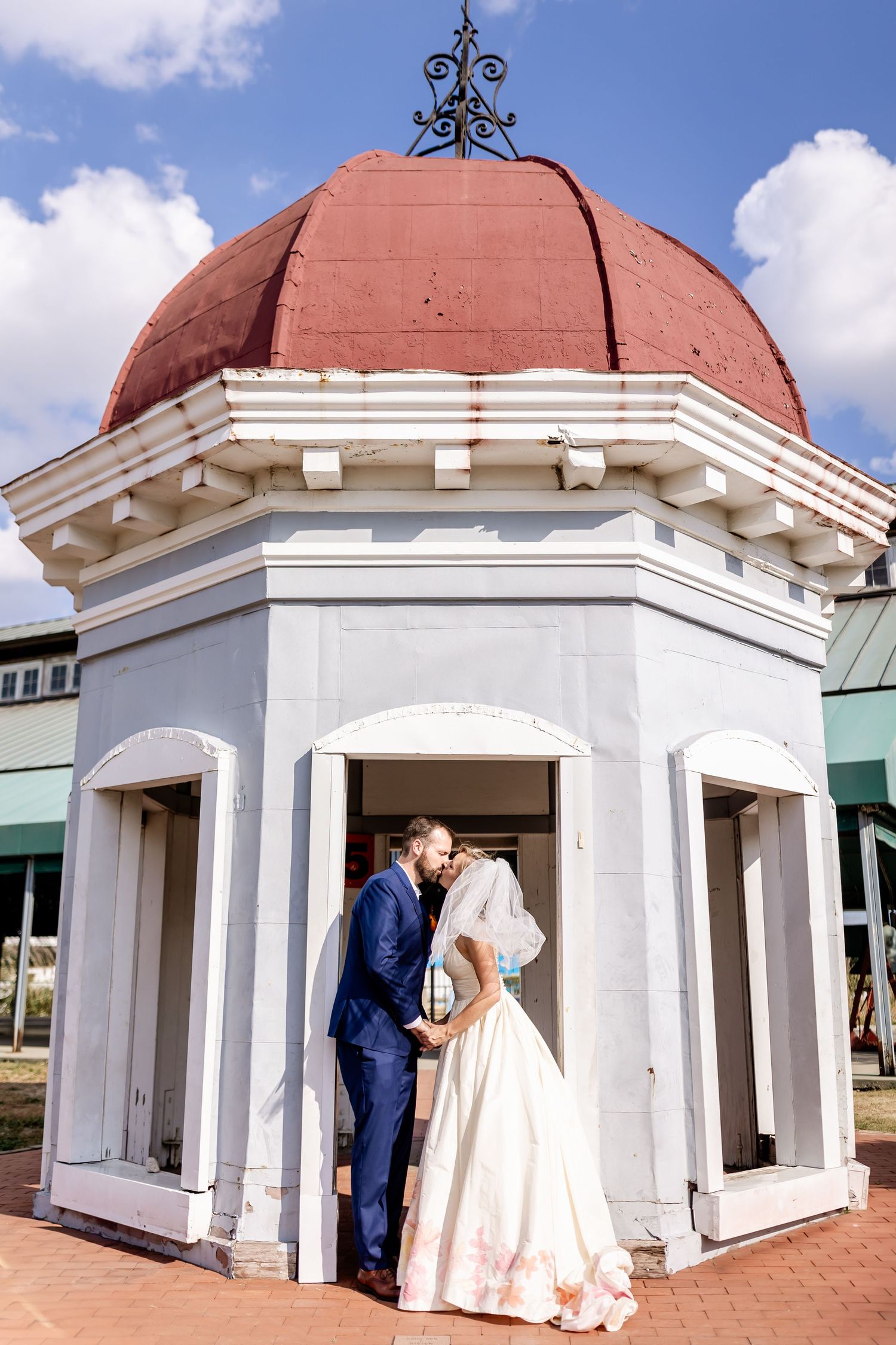 A romantic wedding moment captured under a domed gazebo with terracotta roof and white columns.