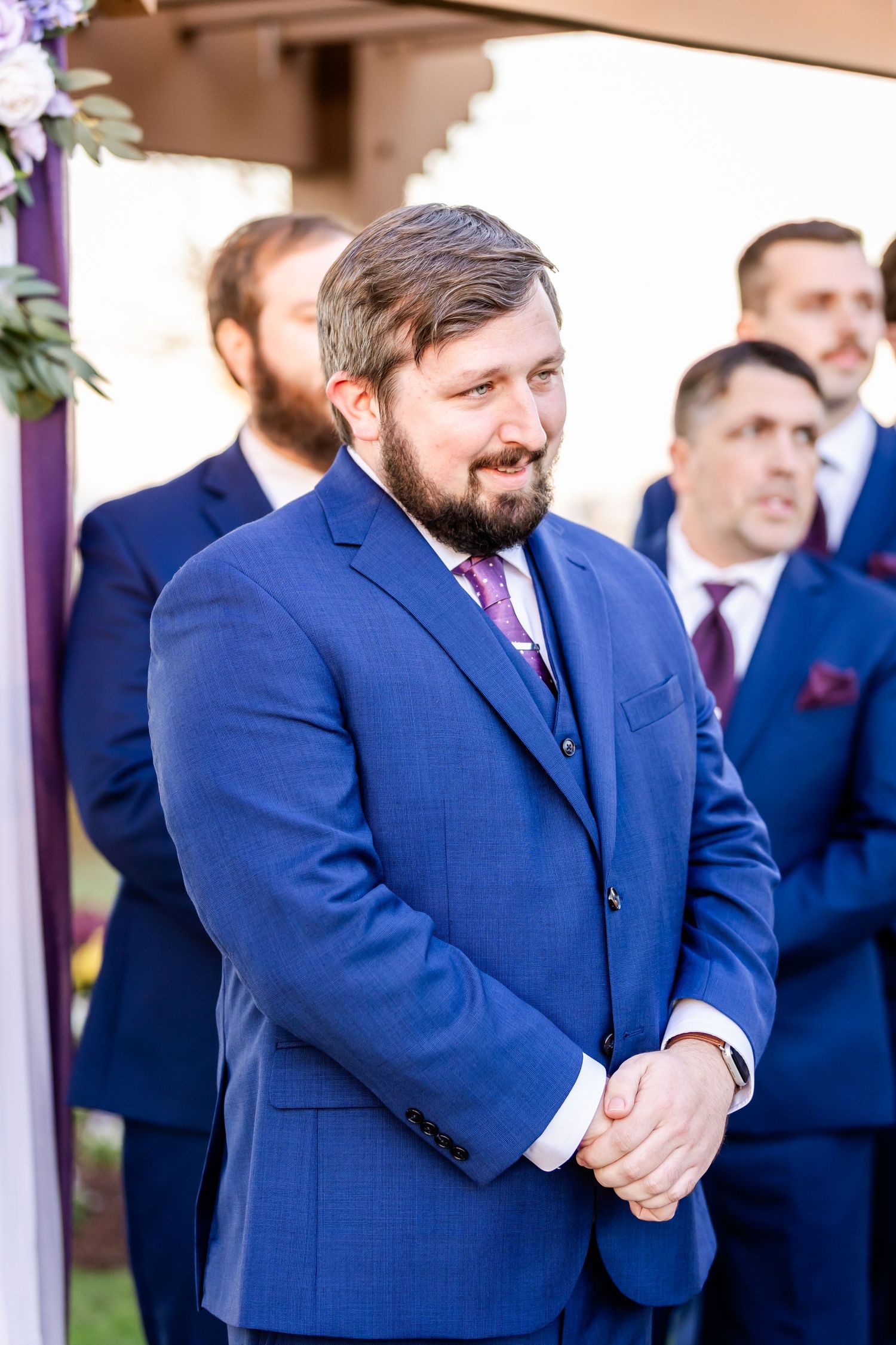 Groomsmen stand in formal attire during outdoor wedding ceremony with man in royal blue suit in focus.