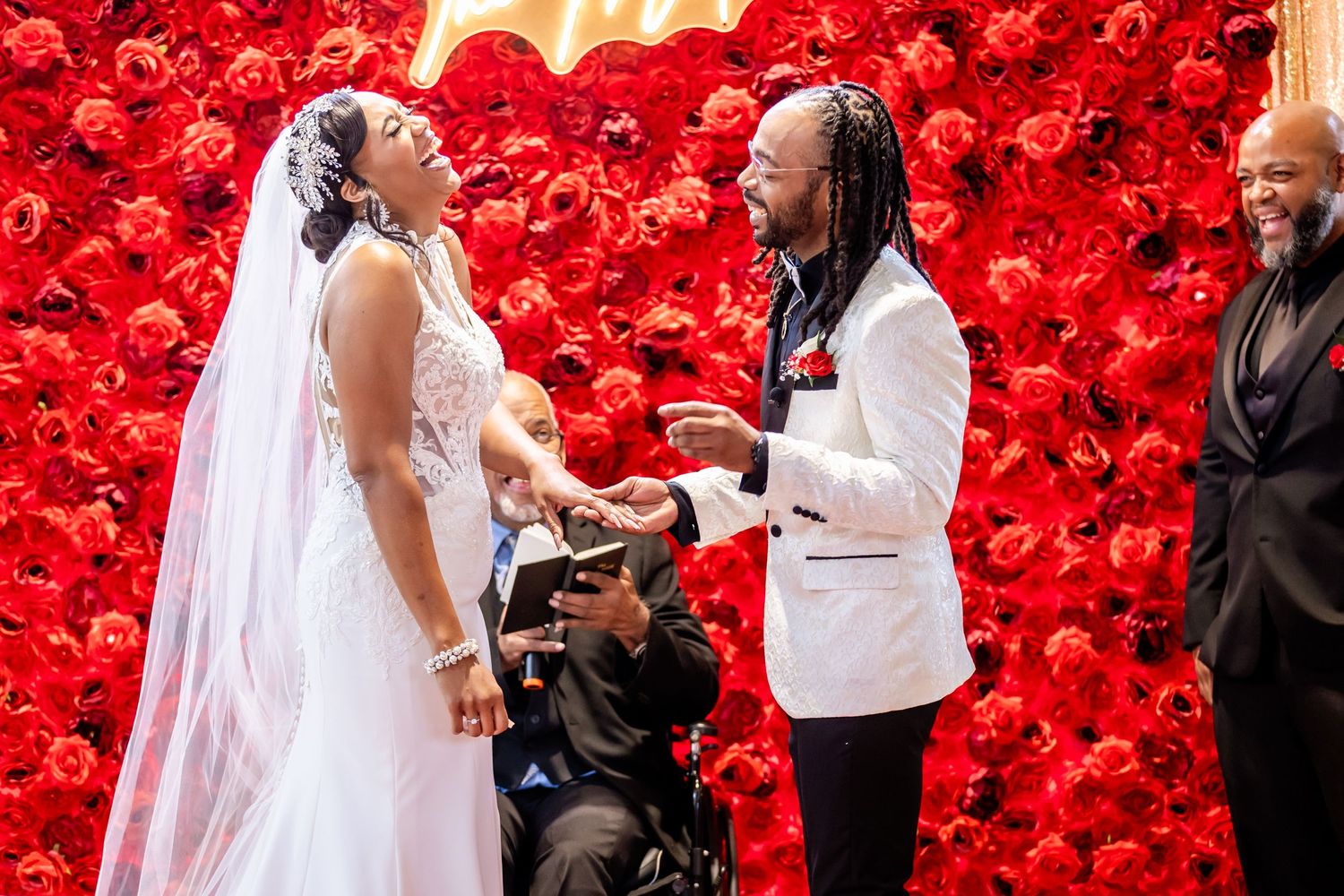 Wedding ceremony exchange of rings in front of vibrant red rose wall.