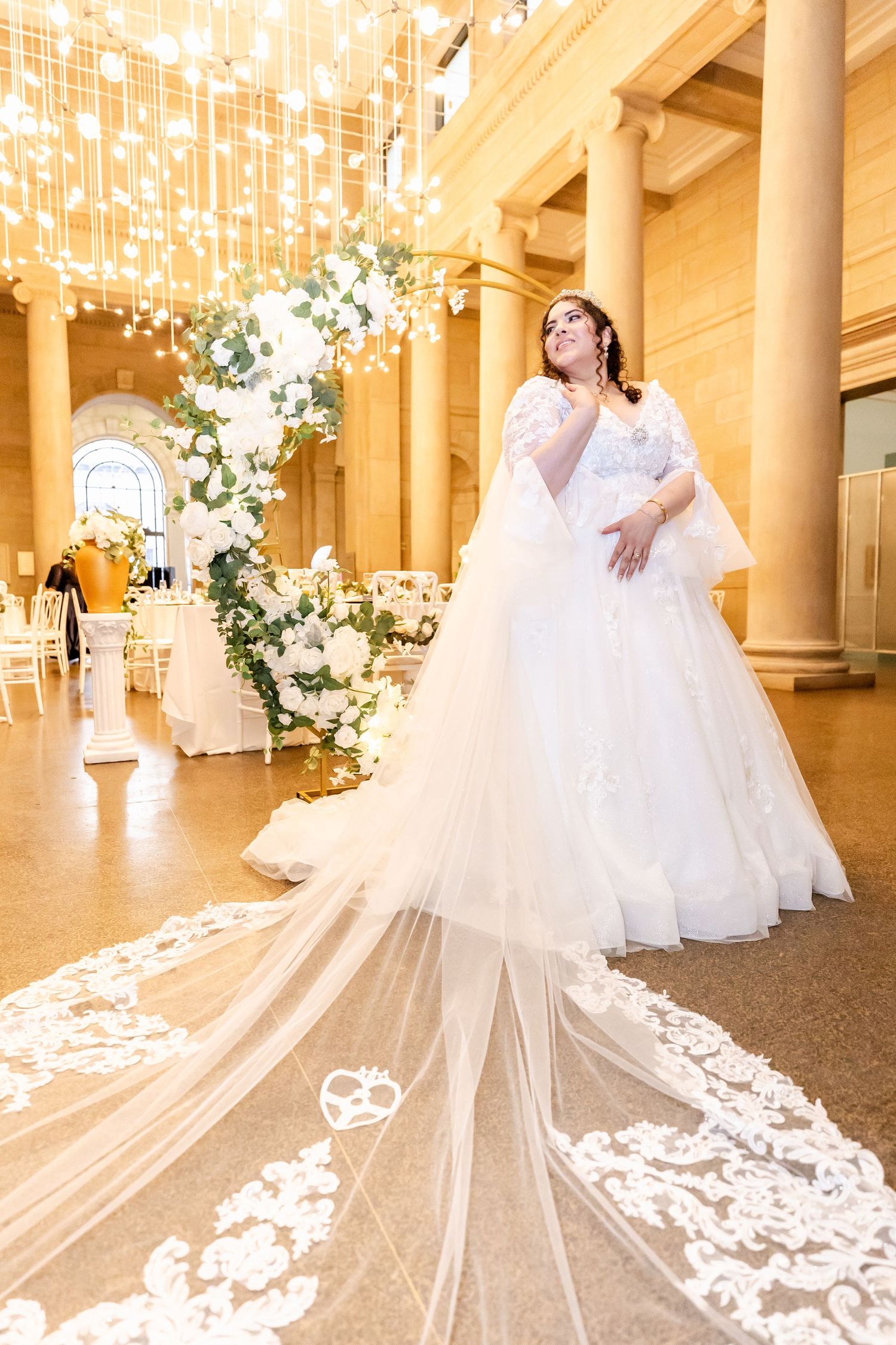 Elegant bride in flowing white gown with long lace veil poses under sparkling chandeliers in grand ballroom setting.