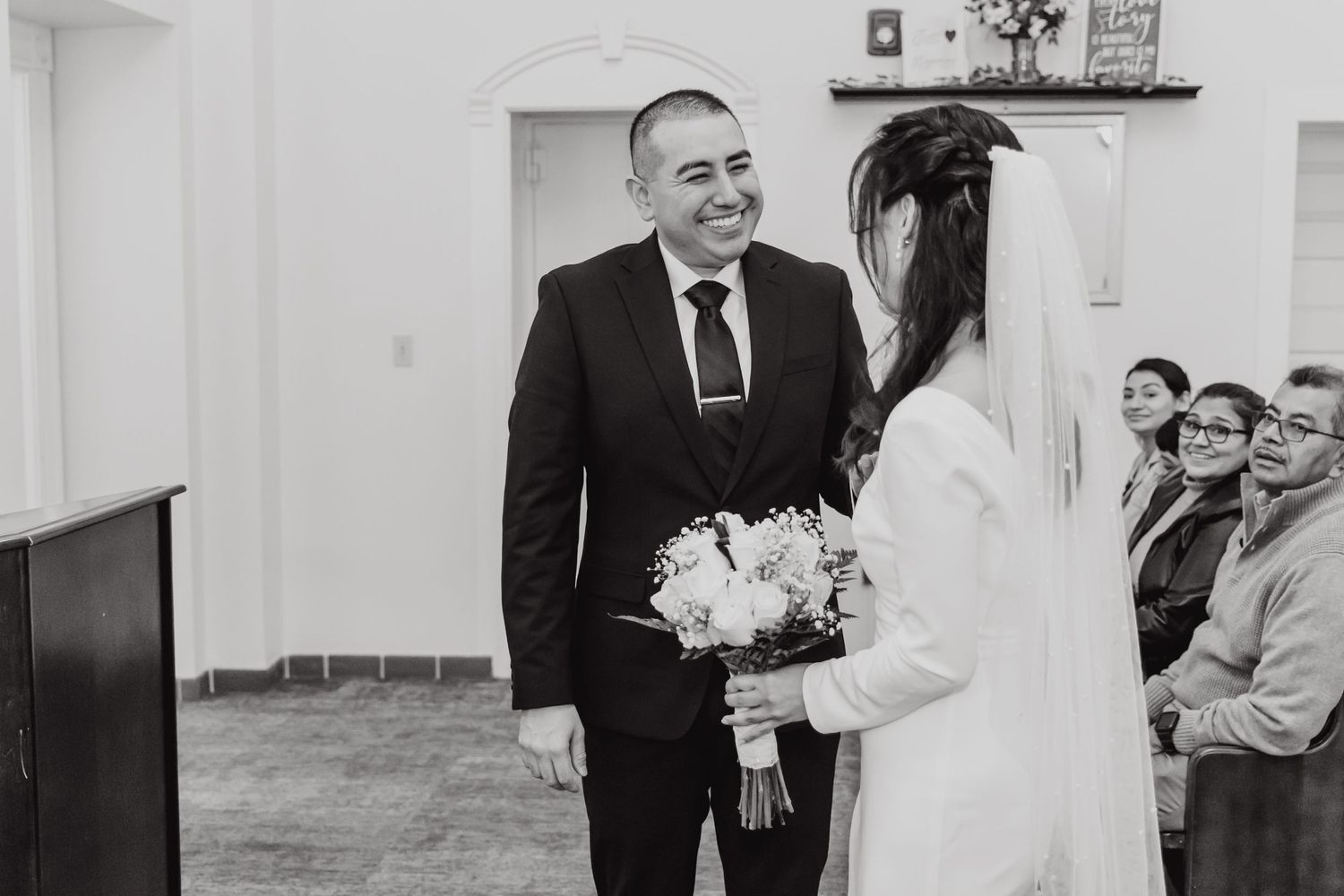An intimate black and white wedding ceremony moment in a simple indoor setting.