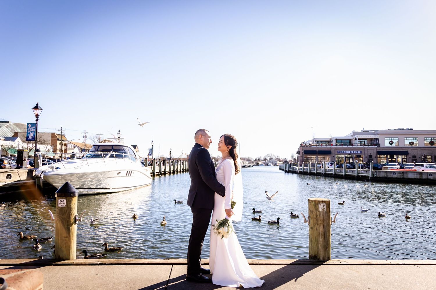 A couple stands on a waterfront dock with boats and seabirds in the background.