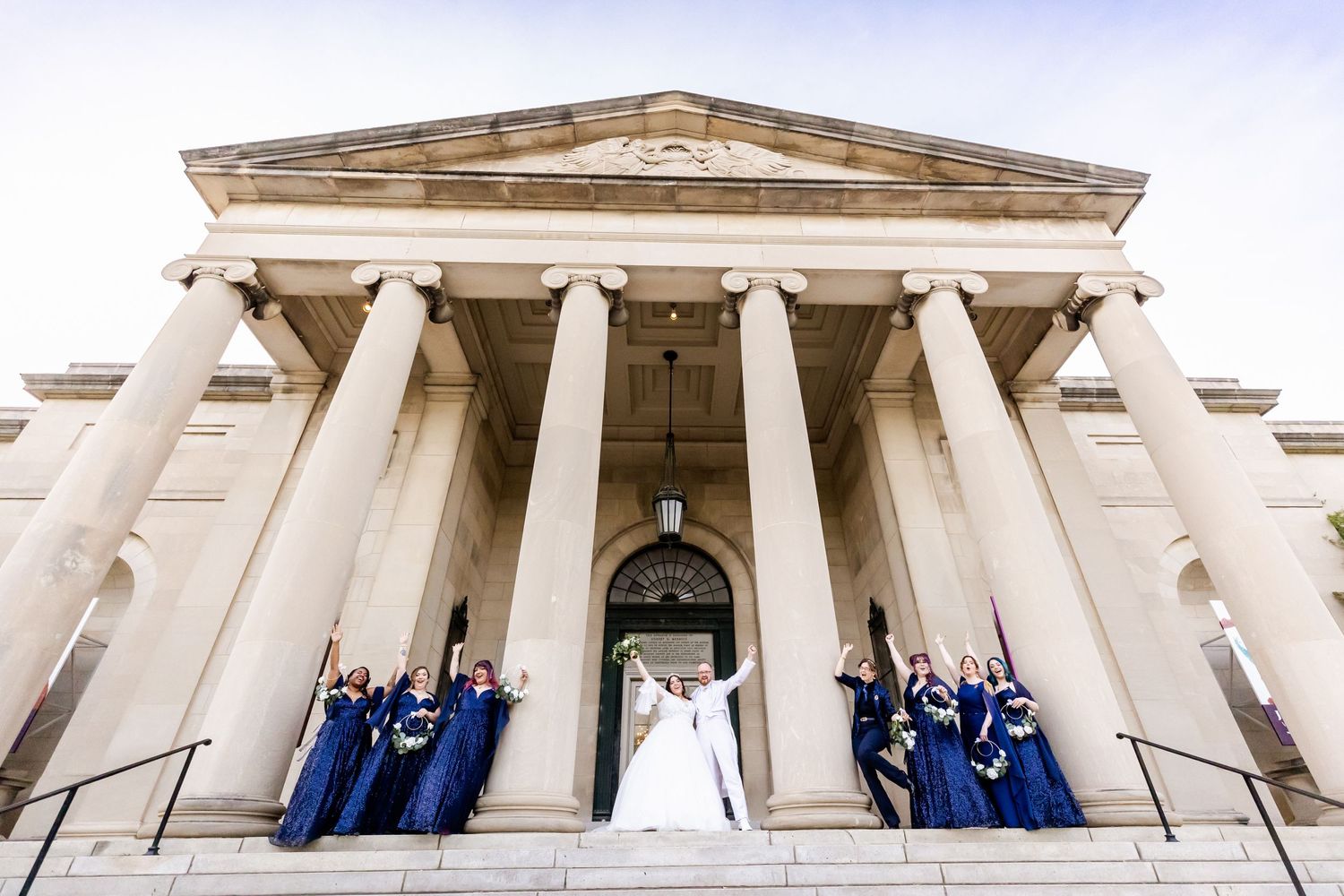 A wedding party poses on the steps of a grand building with classical columns.