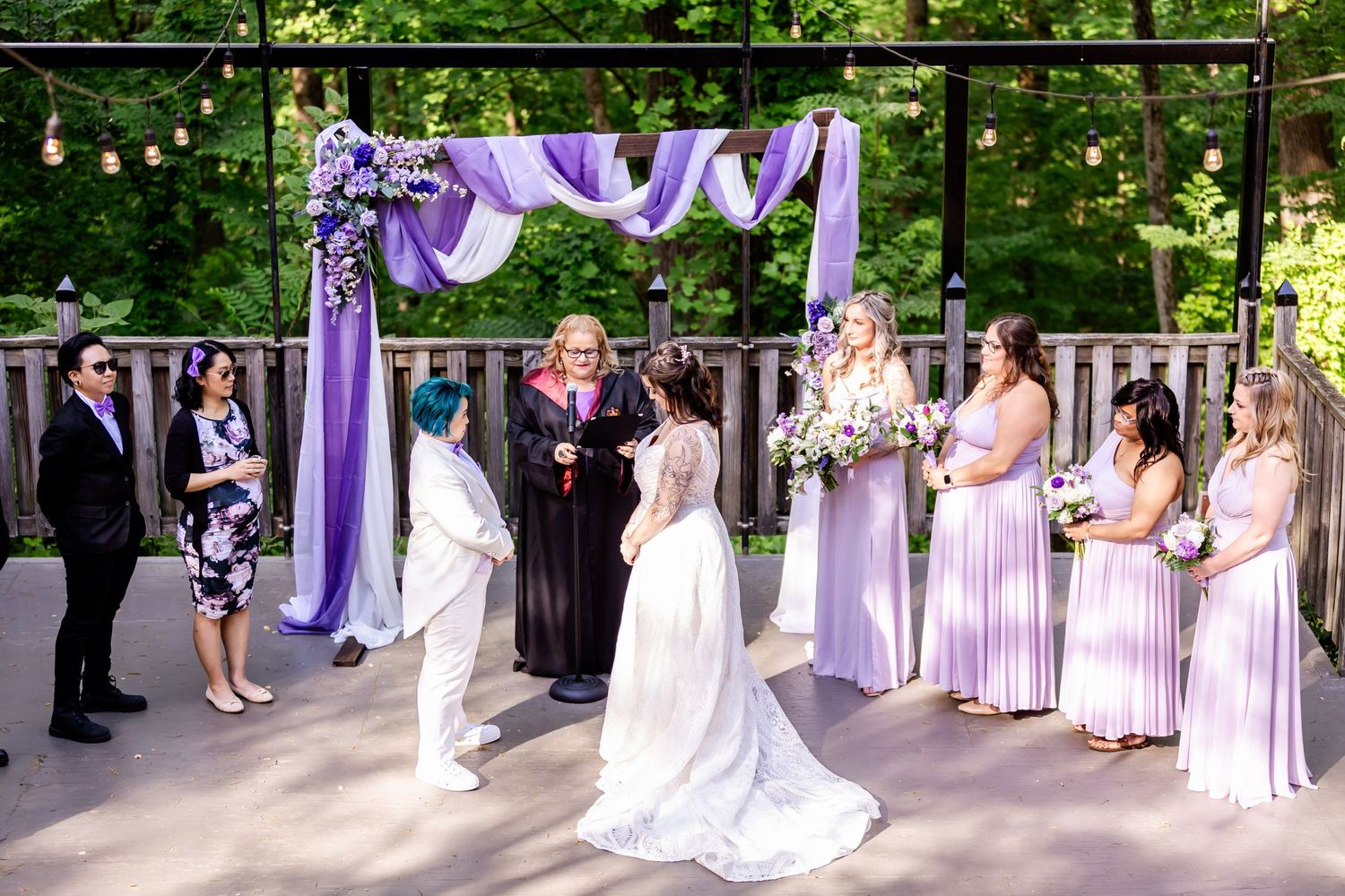 An outdoor wedding ceremony under a purple-draped arch with bridesmaids in lavender dresses.