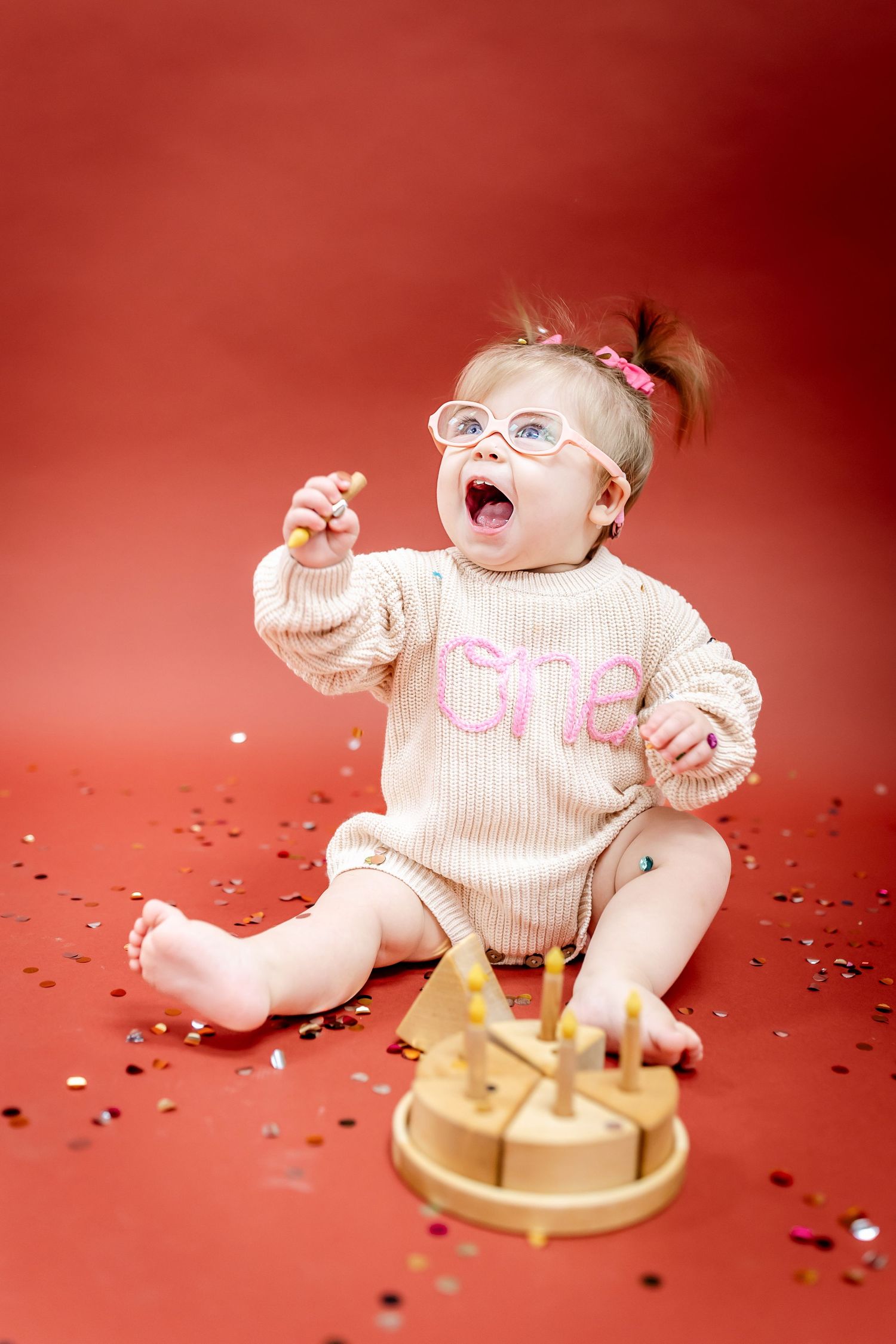 A baby in 'ONE' sweater plays with confetti and gold cake on red background.
