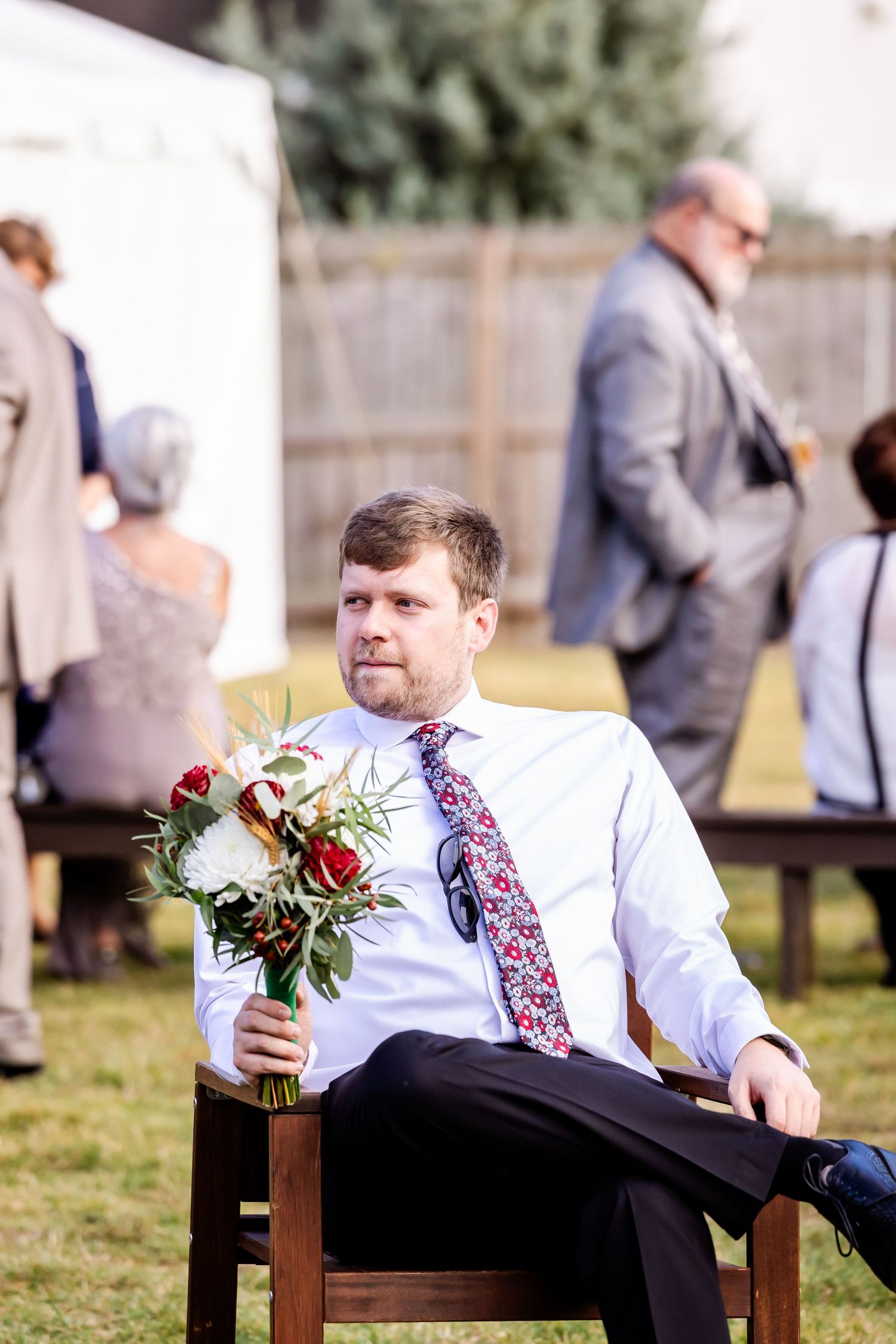 Groomsmen in white shirts with embroidered details relax on wooden chairs while holding red rose boutonnieres.