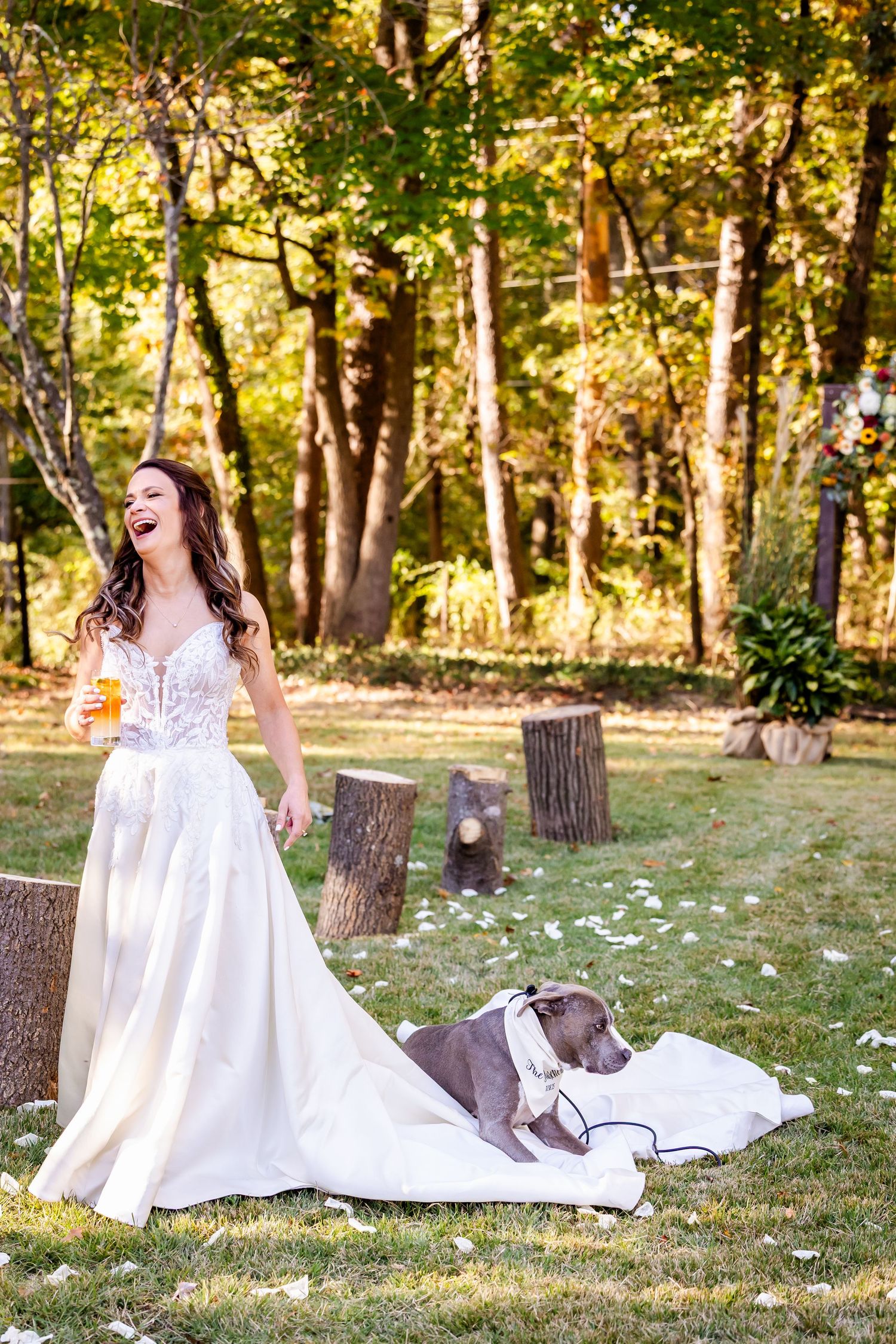 A white wedding dress spreads across green grass in a sunlit wooded setting.