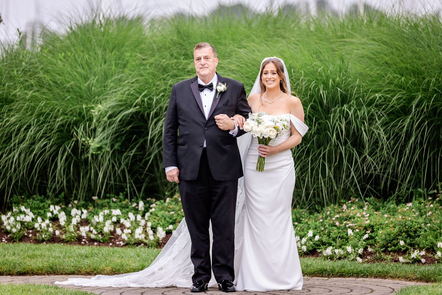 A couple in wedding attire poses for a portrait in front of tall ornamental grasses and white flowers.