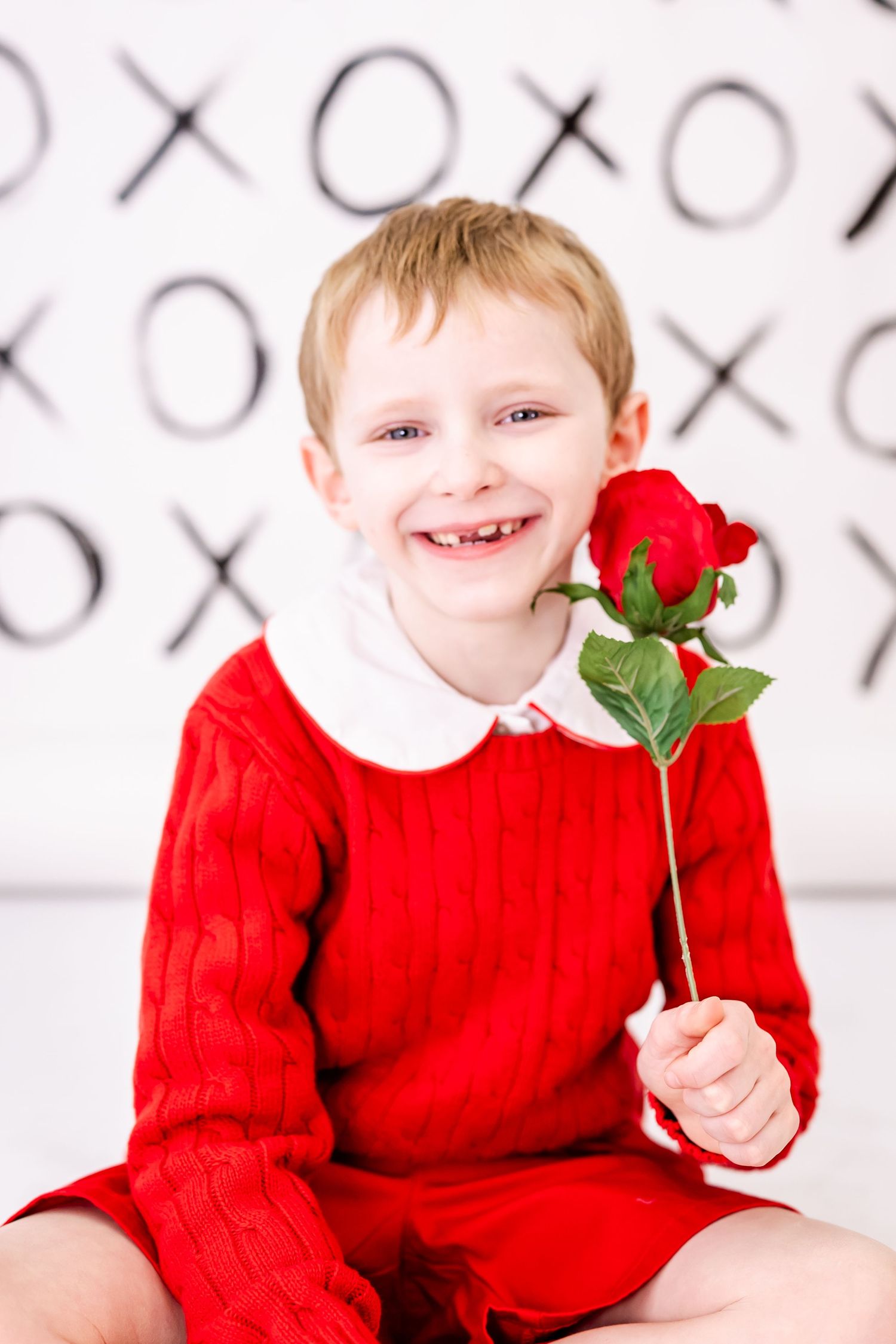 A cheerful smile while holding a red rose against a patterned XOXO background.