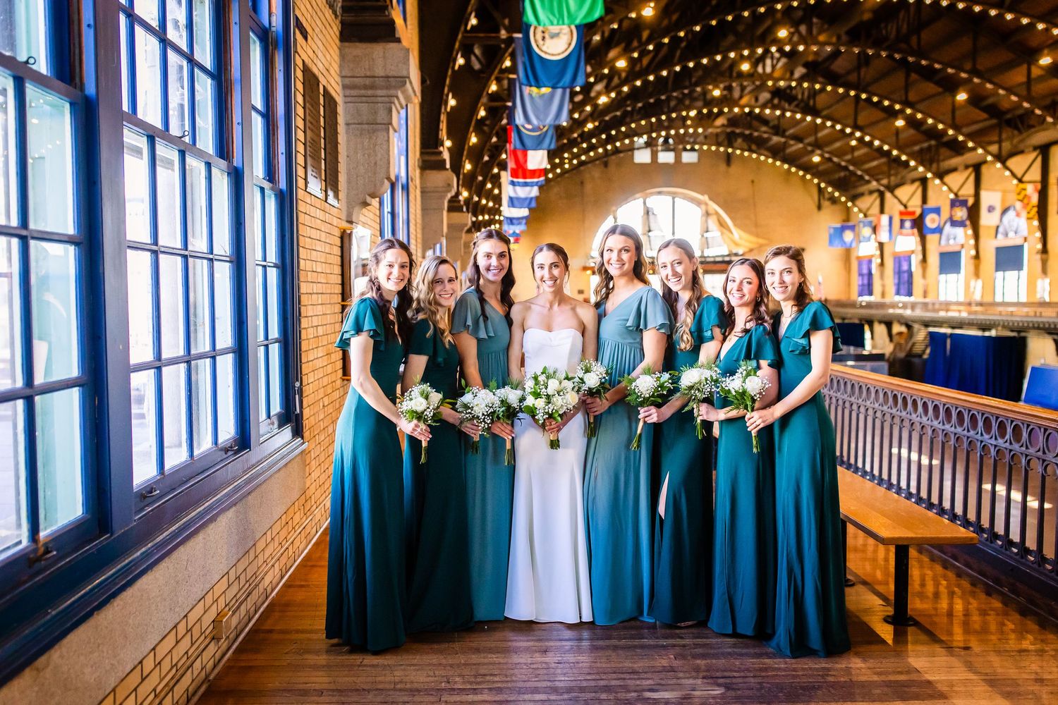 Wedding party poses in elegant teal bridesmaid dresses along a rustic wooden balcony with twinkling lights overhead.