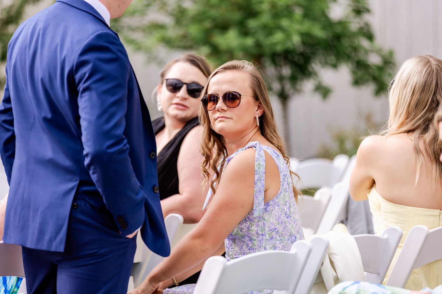 Wedding guests in summer attire seated in white chairs at an outdoor ceremony.