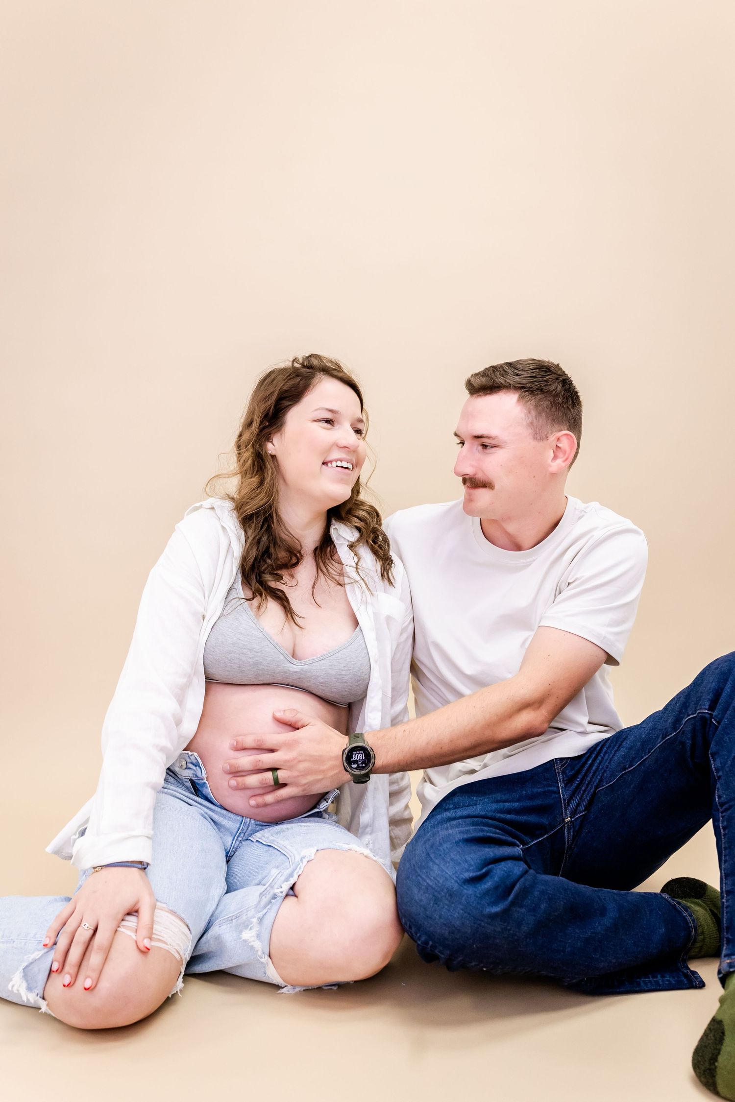 A couple sitting on the floor in casual clothing during a maternity photoshoot against beige background.