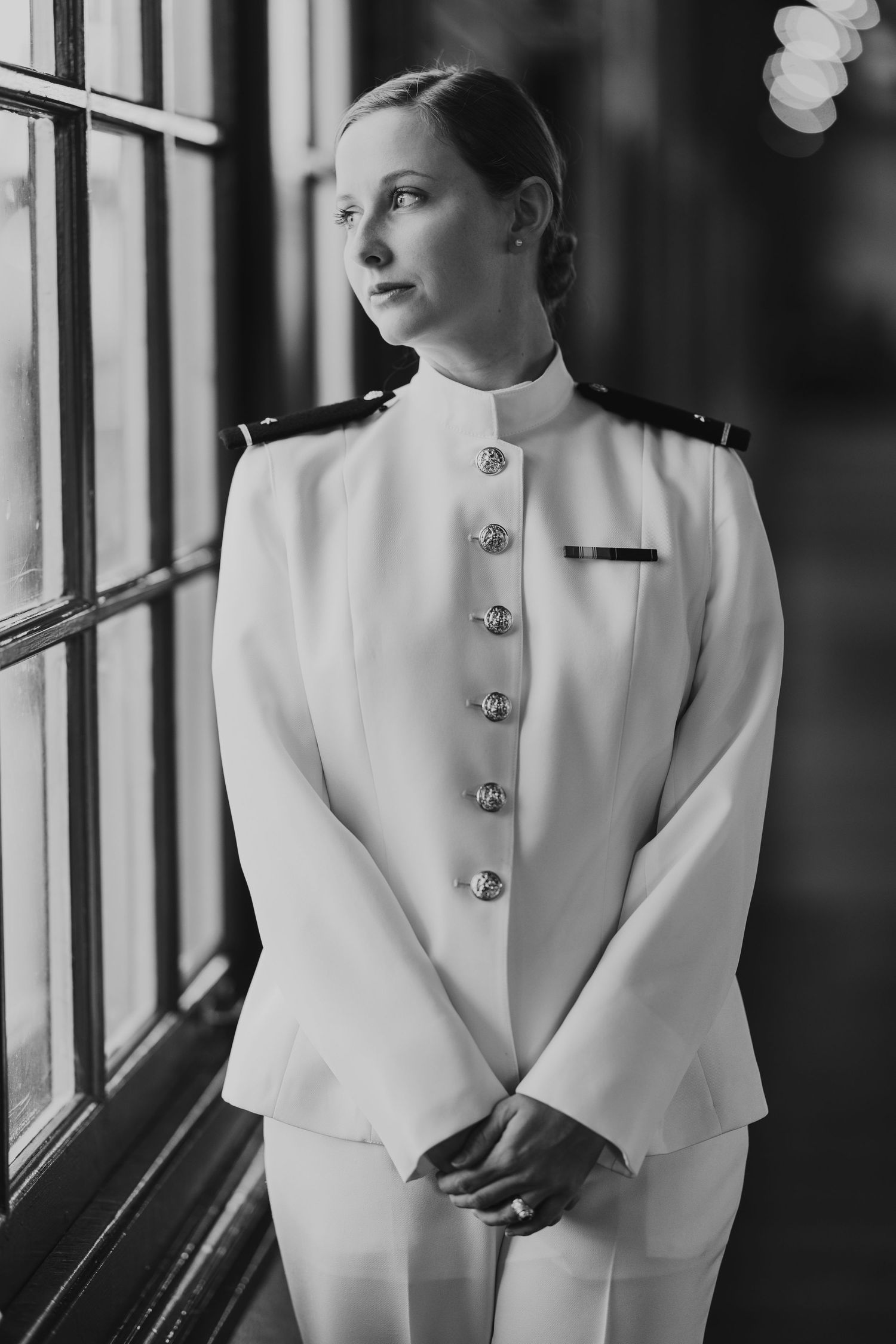 Naval officer in white dress uniform poses by window in black and white portrait.
