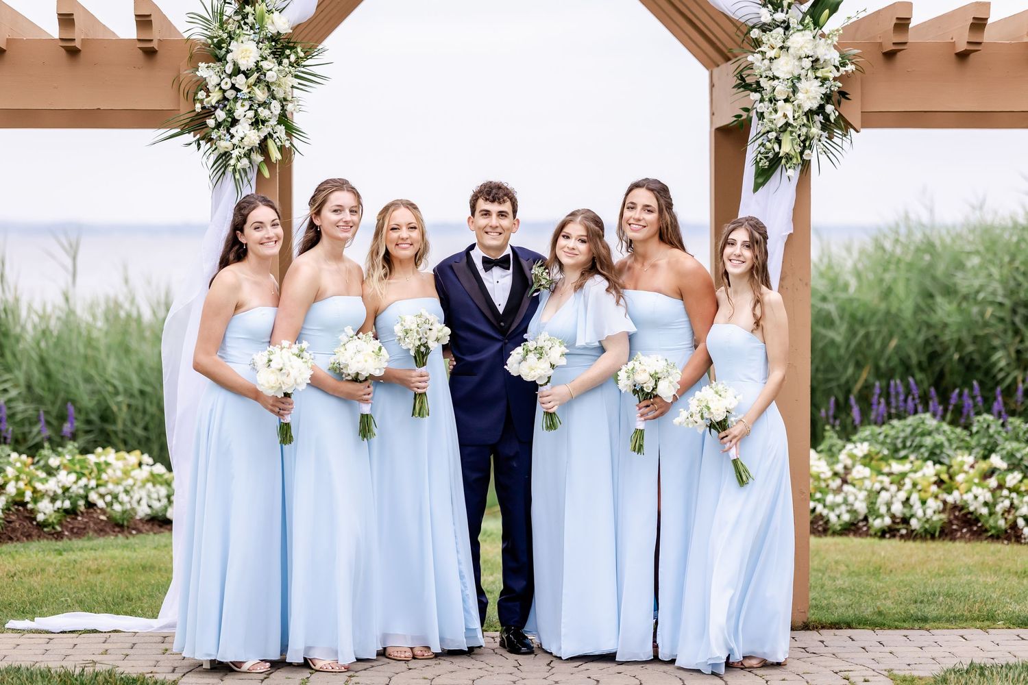 Wedding party poses under floral-decorated pergola with bridesmaids wearing light blue dresses.