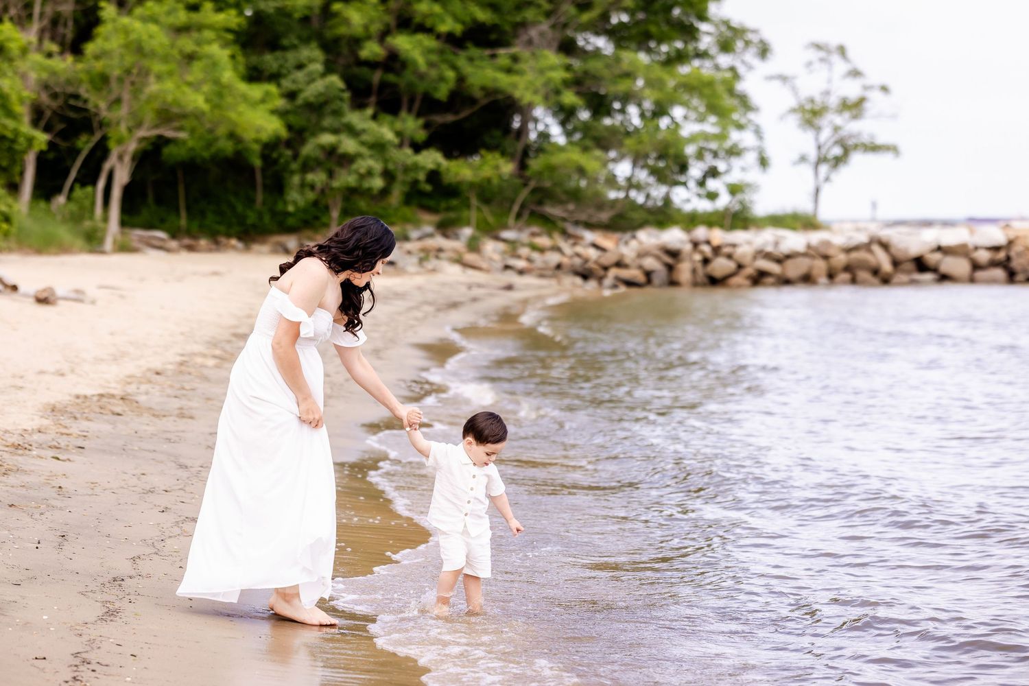 A woman holds a child's hand while walking along a rocky beach shoreline in white dresses.
