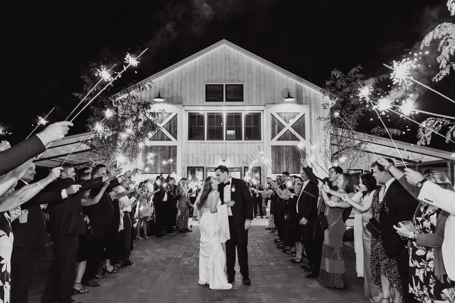 Wedding guests hold sparklers creating a tunnel of light for a couple outside a rustic white barn at night.