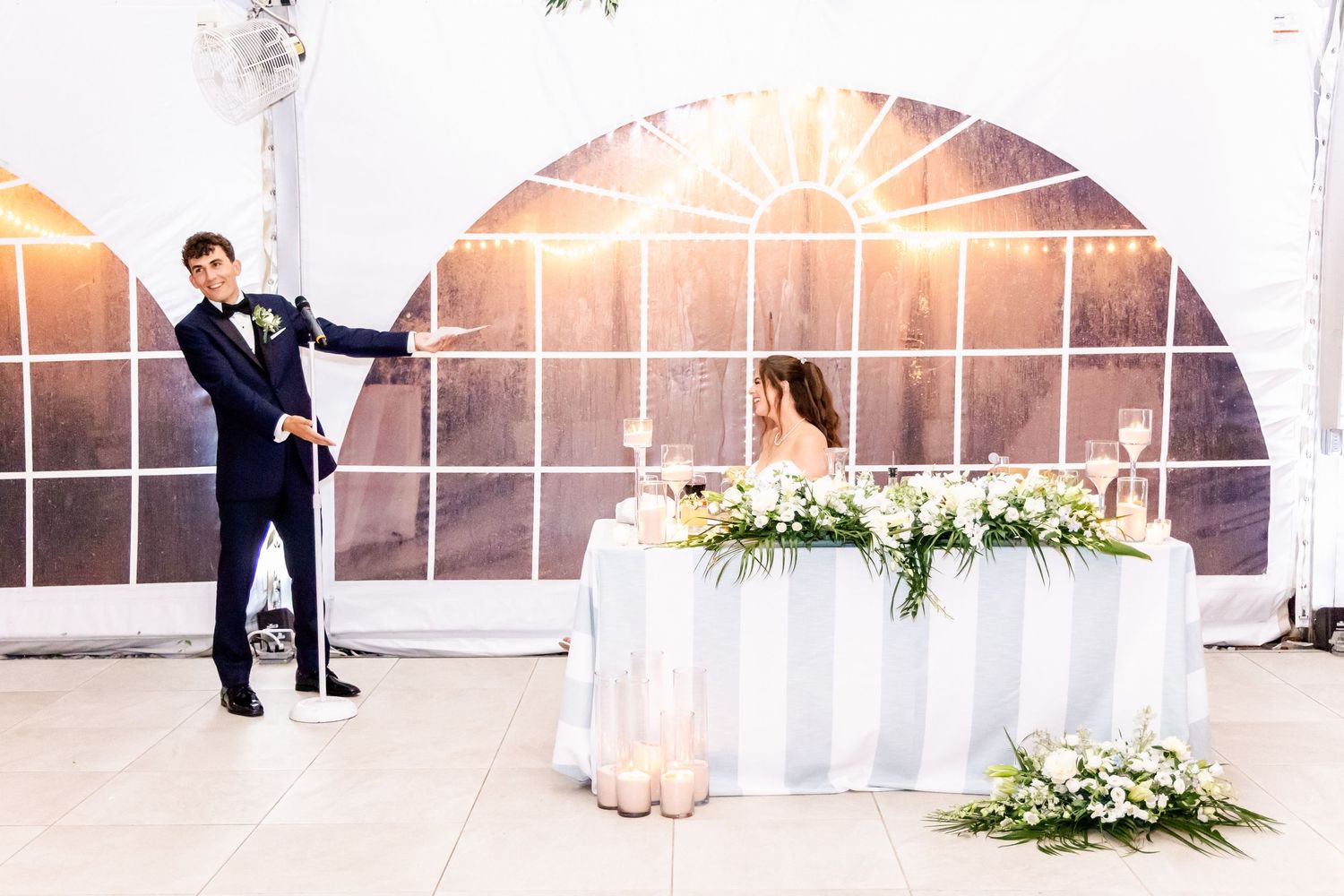 Wedding reception toasts being made in front of a decorated head table with floral arrangements.