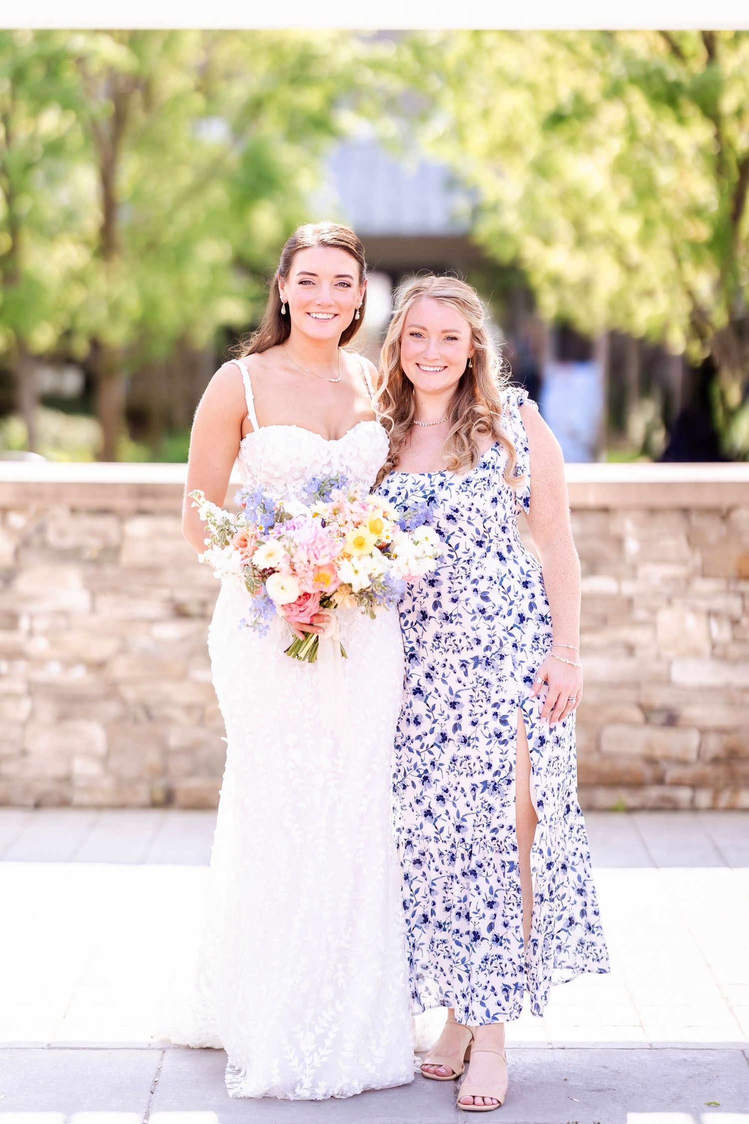 Two women pose outdoors in formal attire with spring floral bouquet.