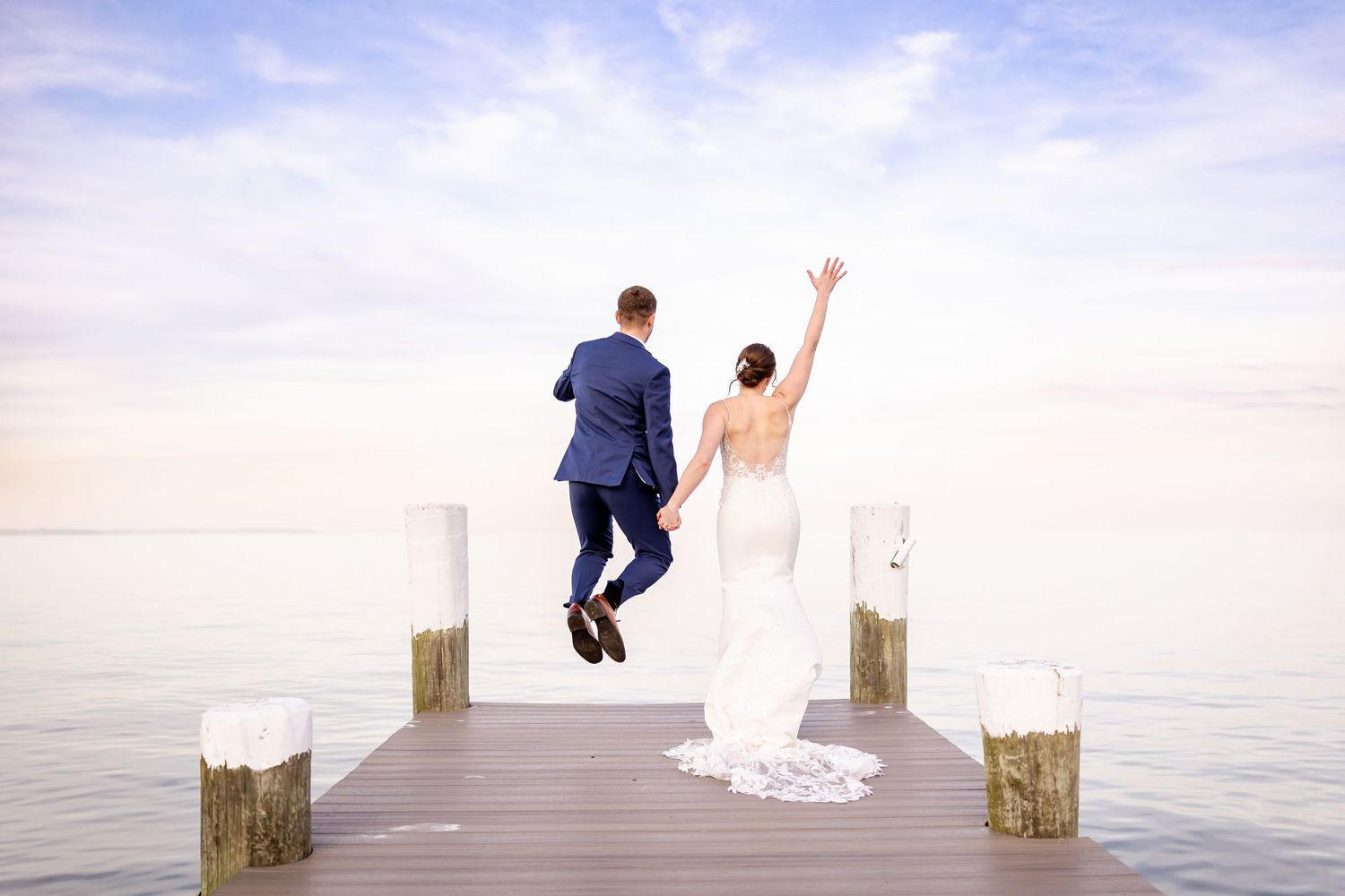 Wedding couple jumps playfully on a wooden pier by the water during sunset photo session.
