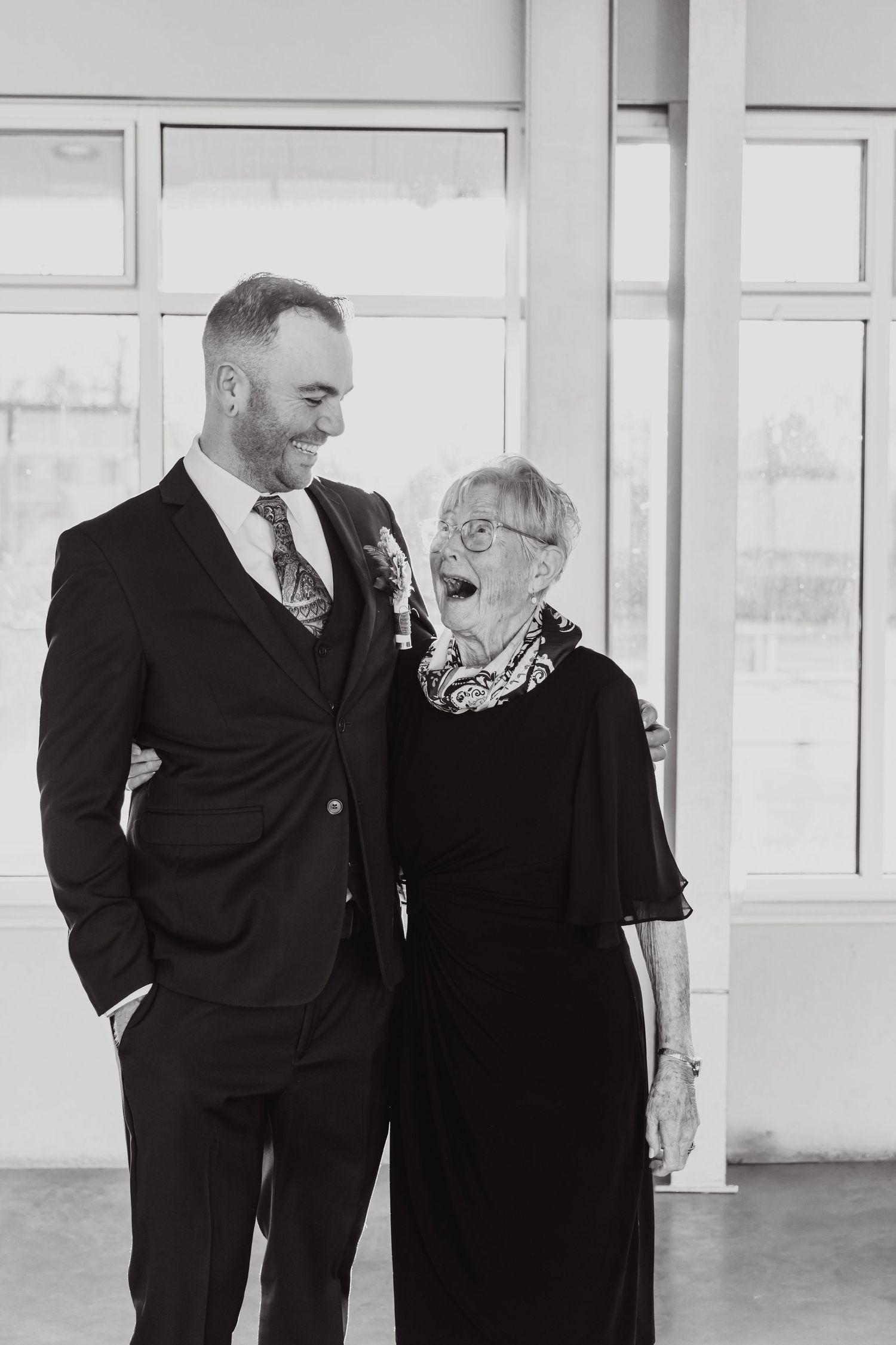 A heartwarming black and white photo showing two people sharing a joyful moment in formal attire by bright windows.