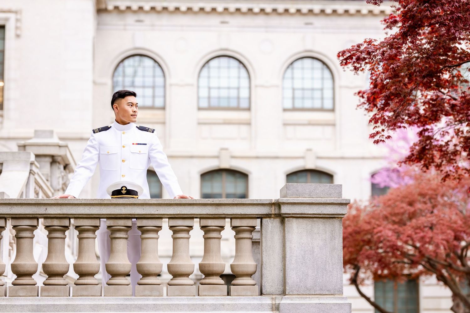 Figure in white military dress uniform poses on ornate balcony with red maple tree and arched windows behind.