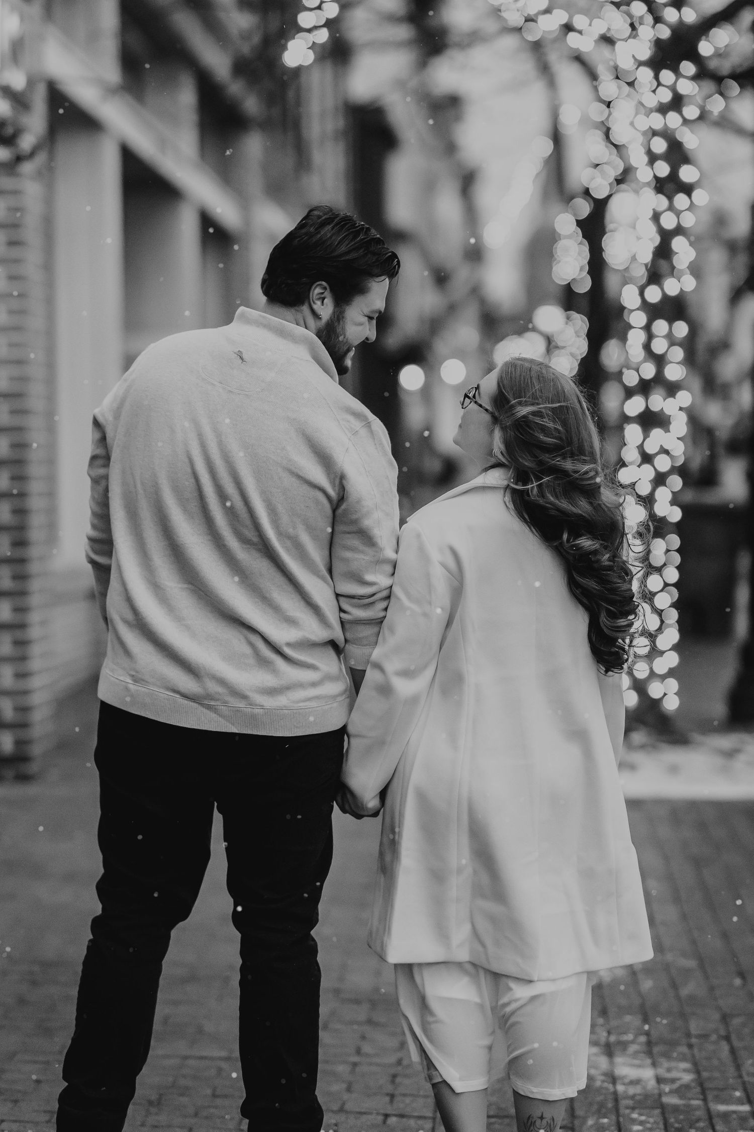 A couple walks together on a cobblestone street at night with twinkling holiday lights in the background.