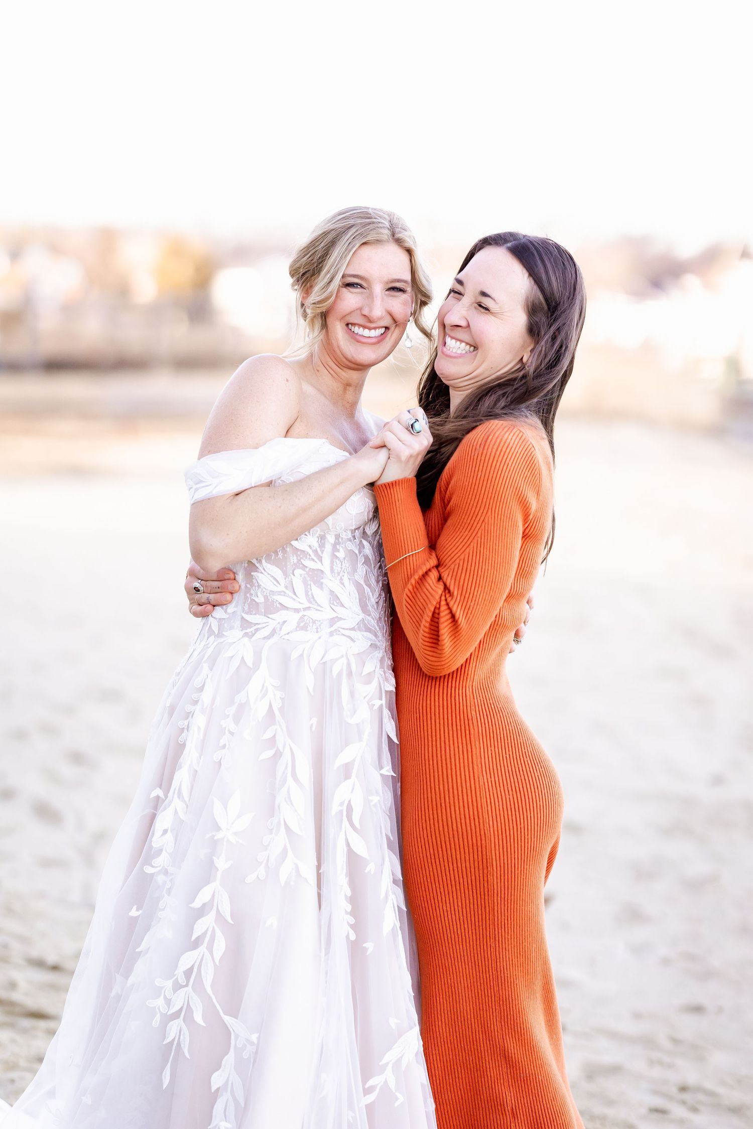 Close friends share a joyful moment on the beach, one in a white gown and one in orange sweater dress.