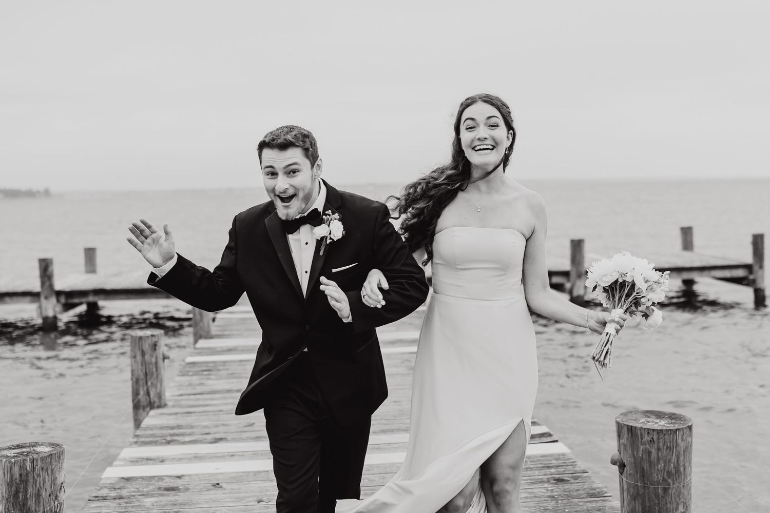 Wedding couple celebrates on a sandy beach with arms outstretched in black and white photography.
